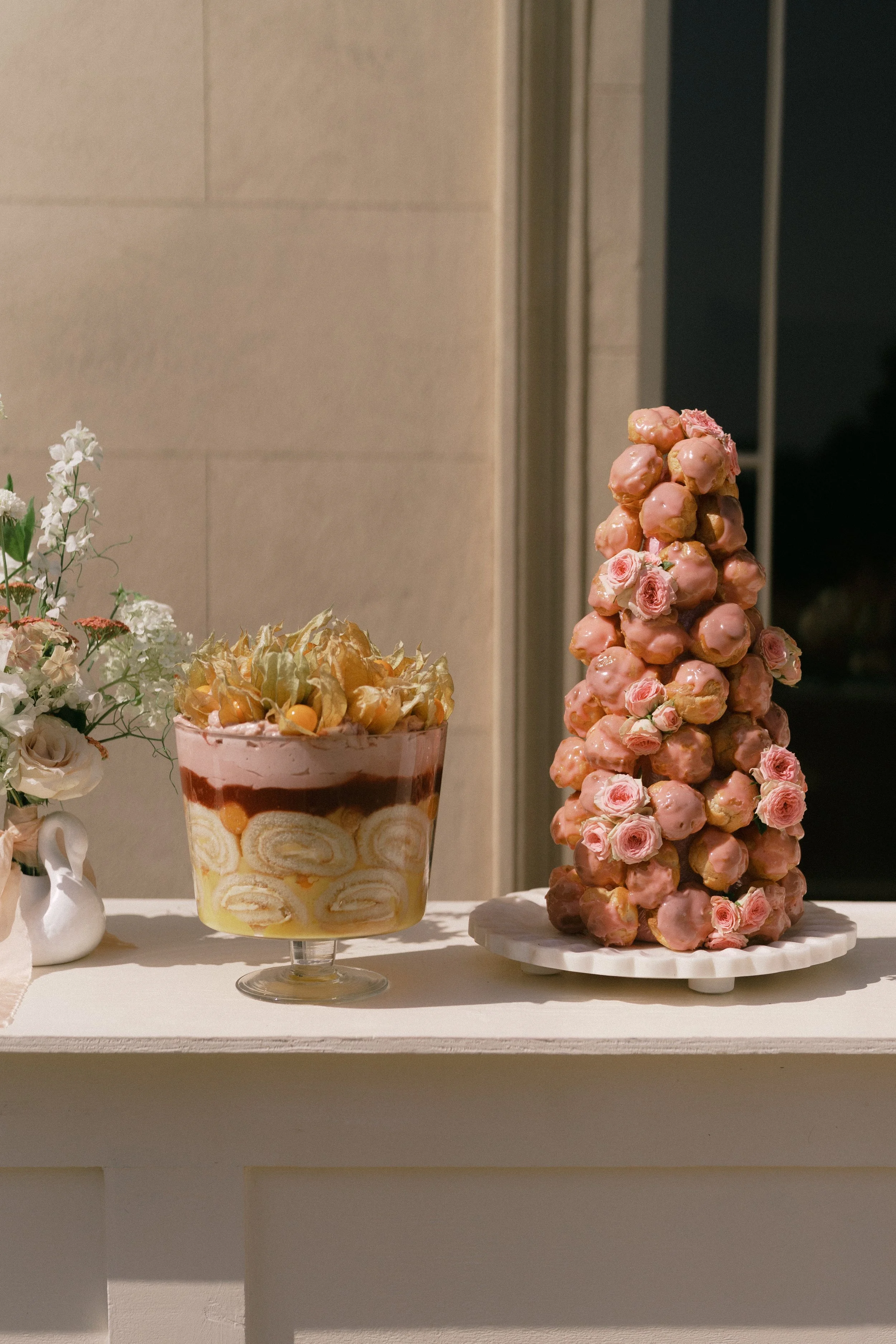Decorative display of desserts and flowers on a white shelf, featuring a tall pink croquembouche topped with pink roses, a layered cake with cream and fruit, and a small white vase with flowers.