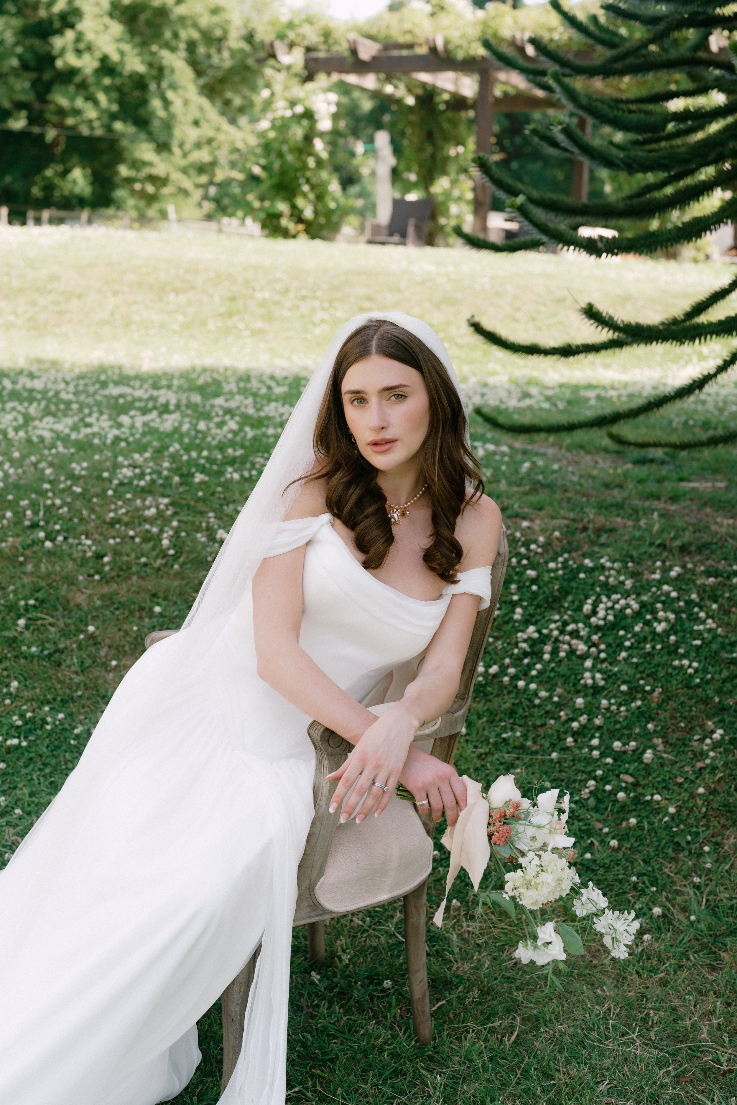 A young woman in a white wedding dress seated on a wooden chair outdoors, holding a bouquet of white flowers, with trees and a green lawn in the background.