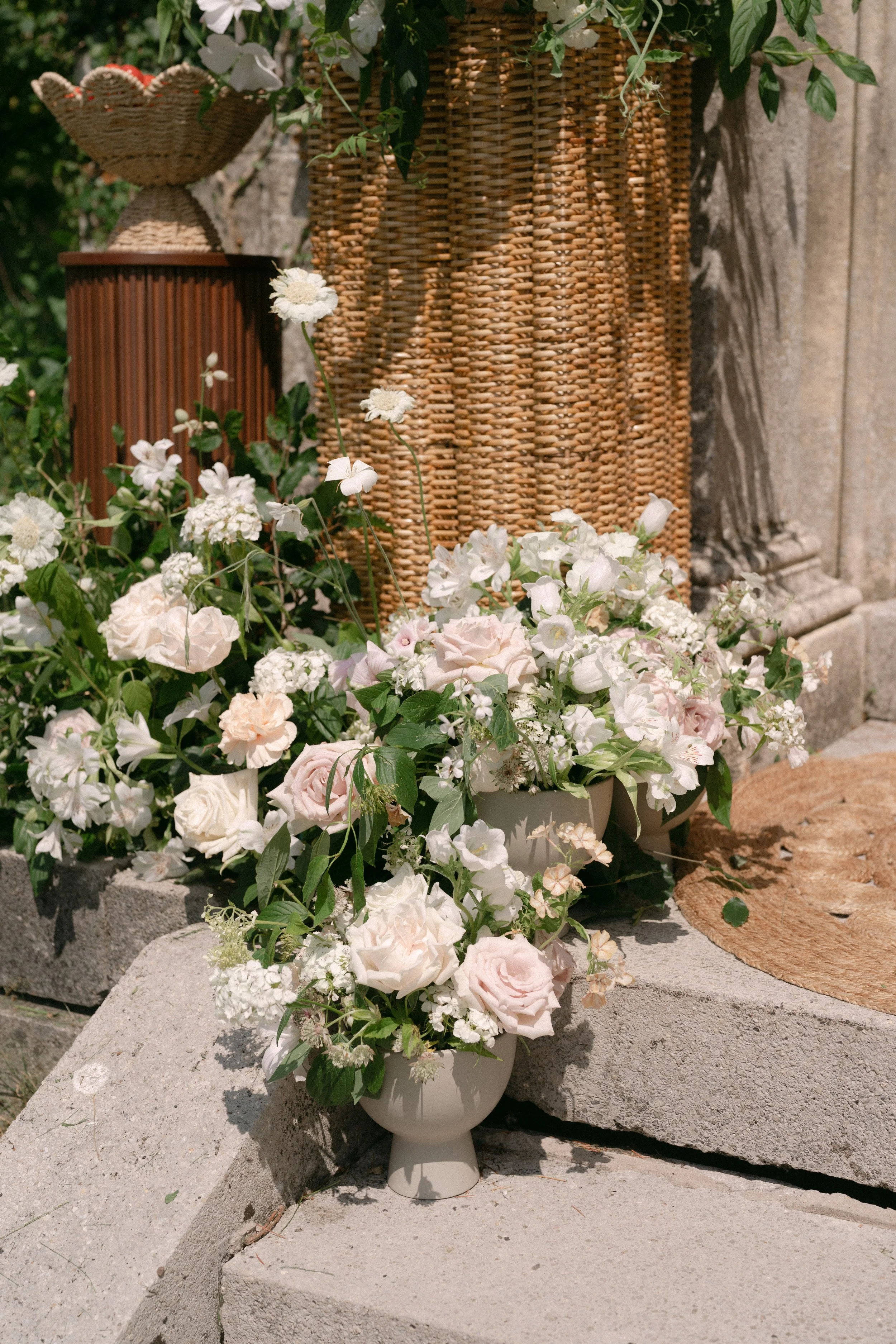 Two white flower arrangements in vases on a stone surface, with wicker baskets and a stone wall in the background.
