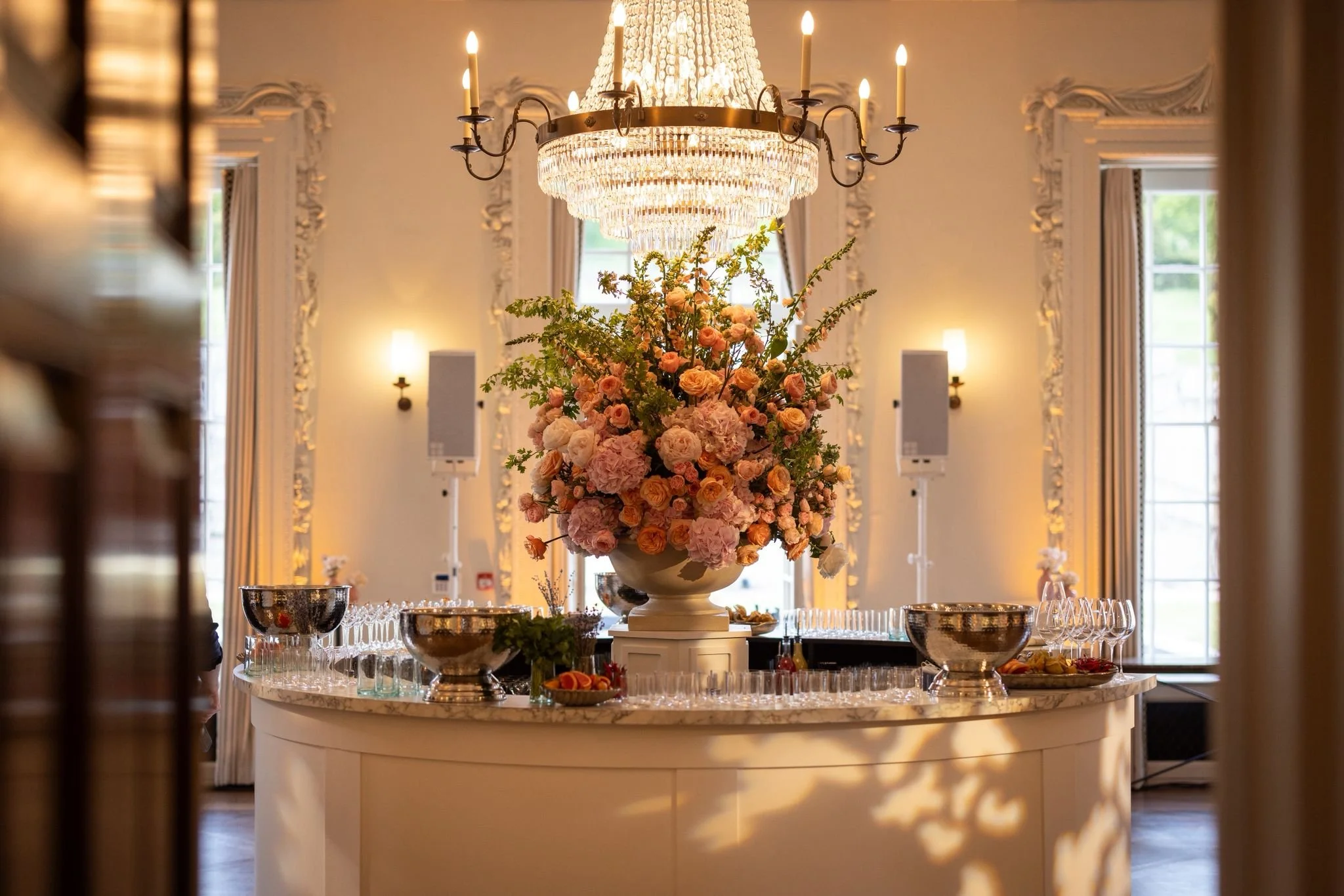 A large floral centerpiece with pink and peach roses, hydrangeas, and greenery on a marble table in a decorated room with a crystal chandelier overhead and windows in the background.
