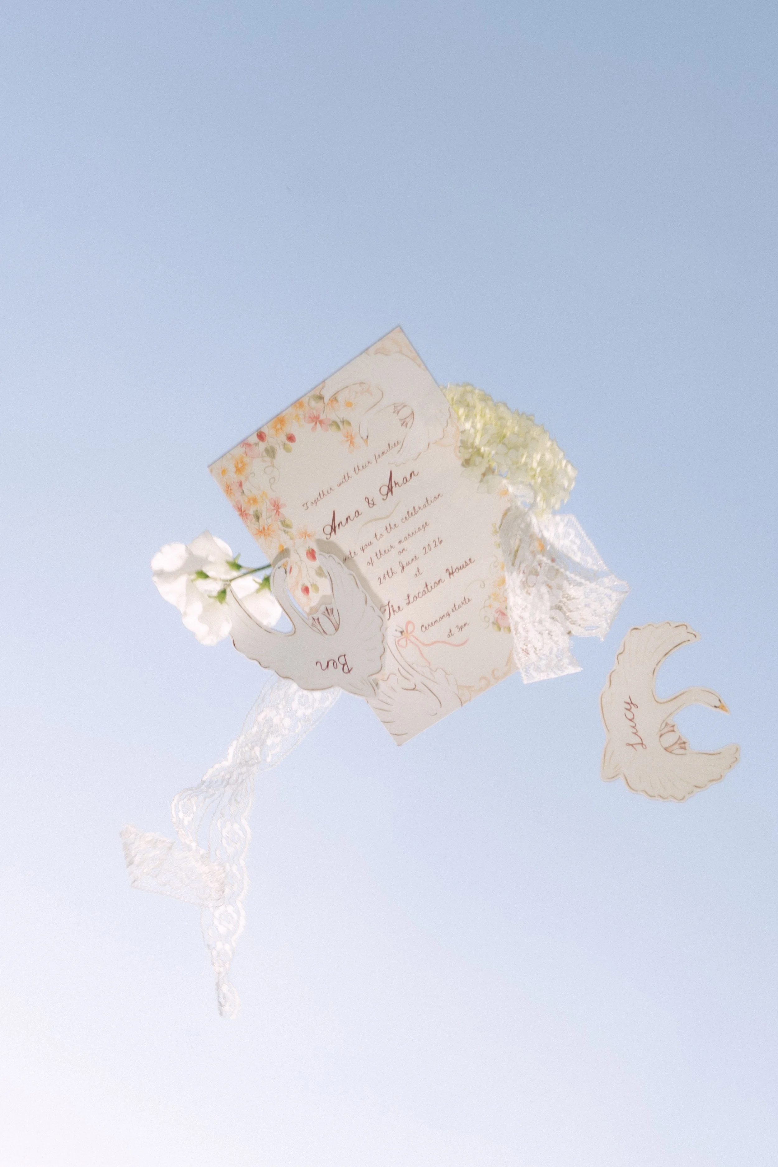 Decorative wedding invitation hanging against a clear blue sky with lace ribbons and white flowers.