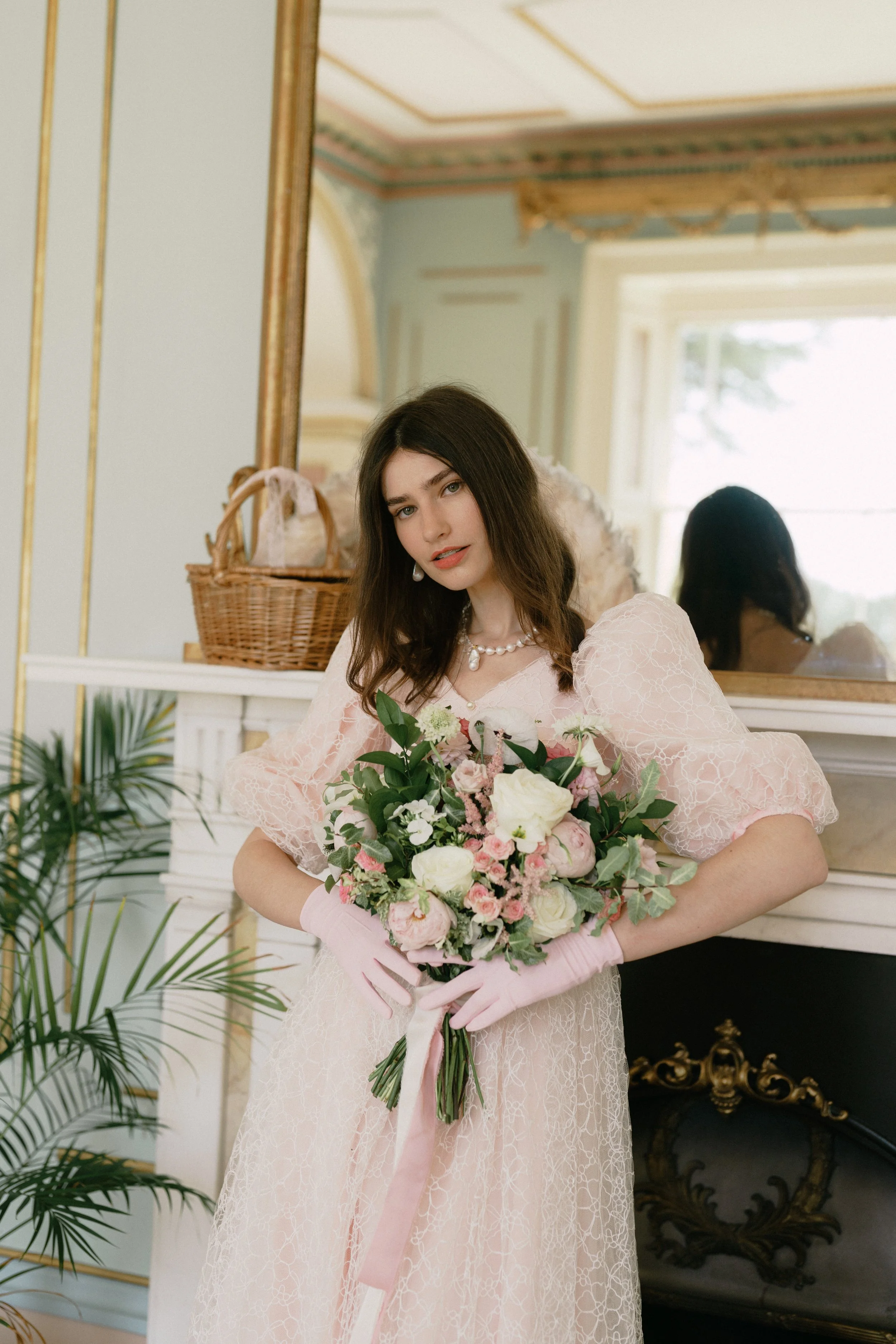 A young woman with dark wavy hair dressed in a vintage pink lace gown holding a large bouquet of white and pink flowers with greenery, standing in an elegant, historic room with ornate decor.