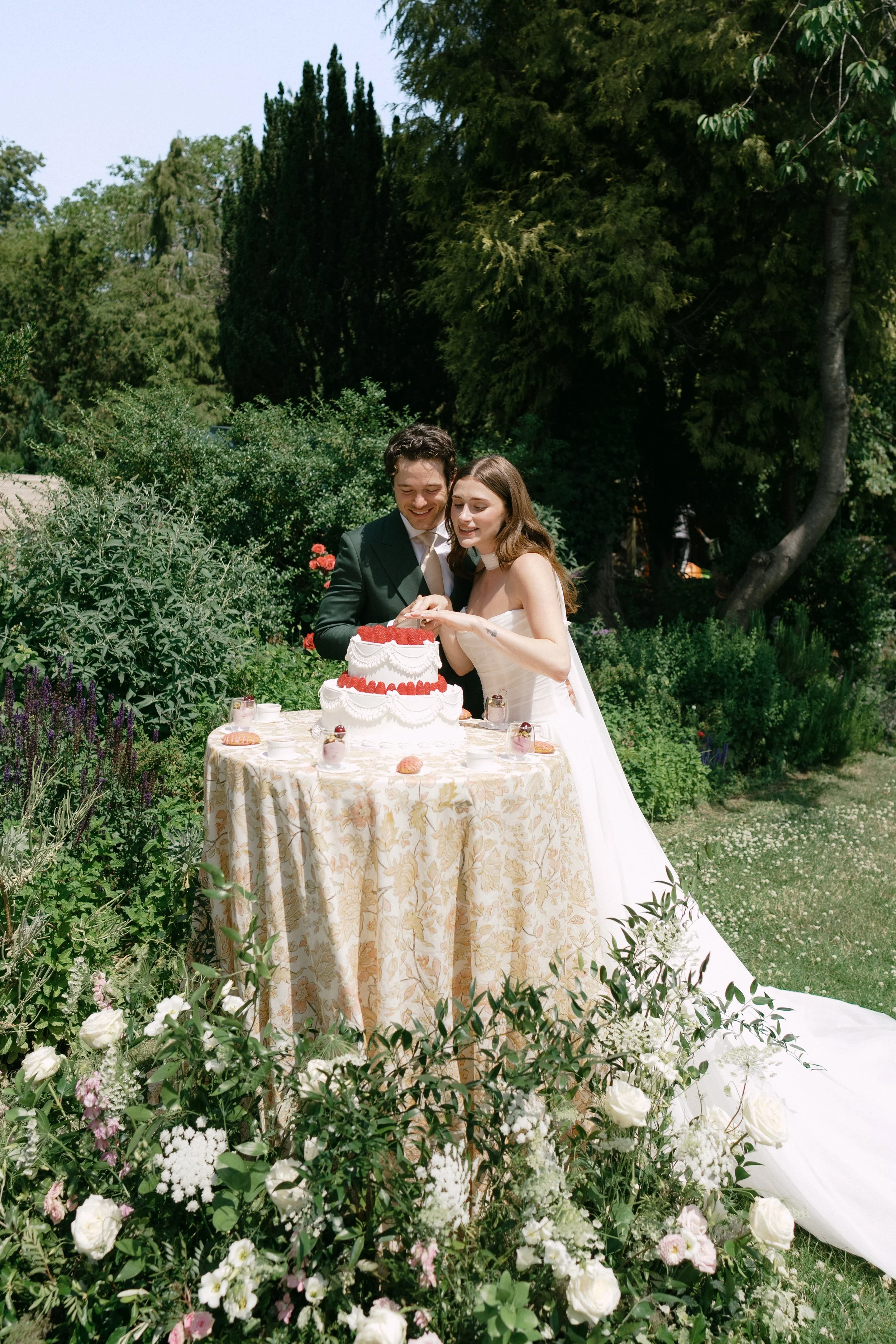 A bride and groom cutting a wedding cake outdoors in a garden surrounded by lush green trees and flowers.