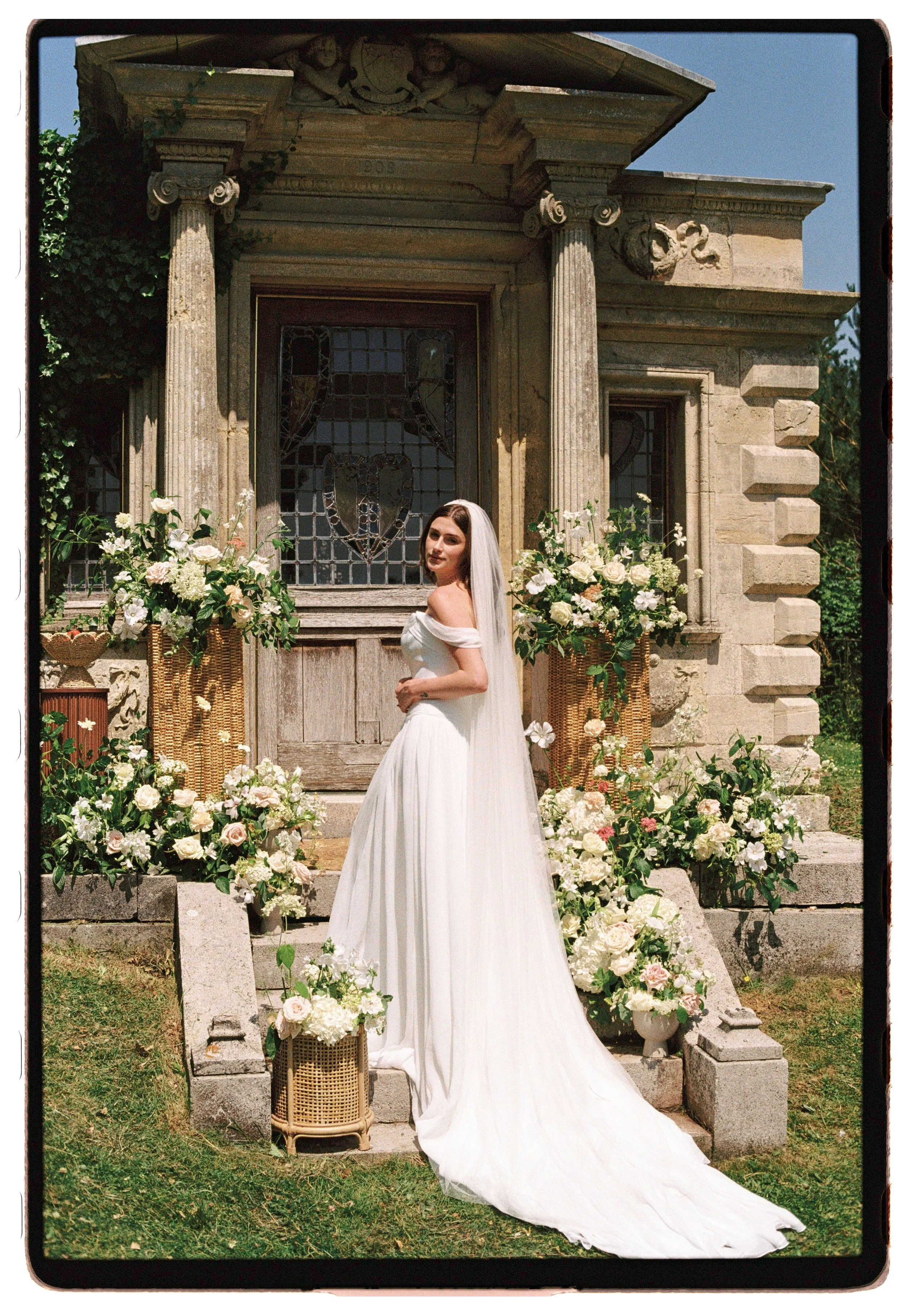 A bride in a white wedding gown standing outside in front of a stone building with stained glass window, surrounded by large floral arrangements on either side and at her feet.