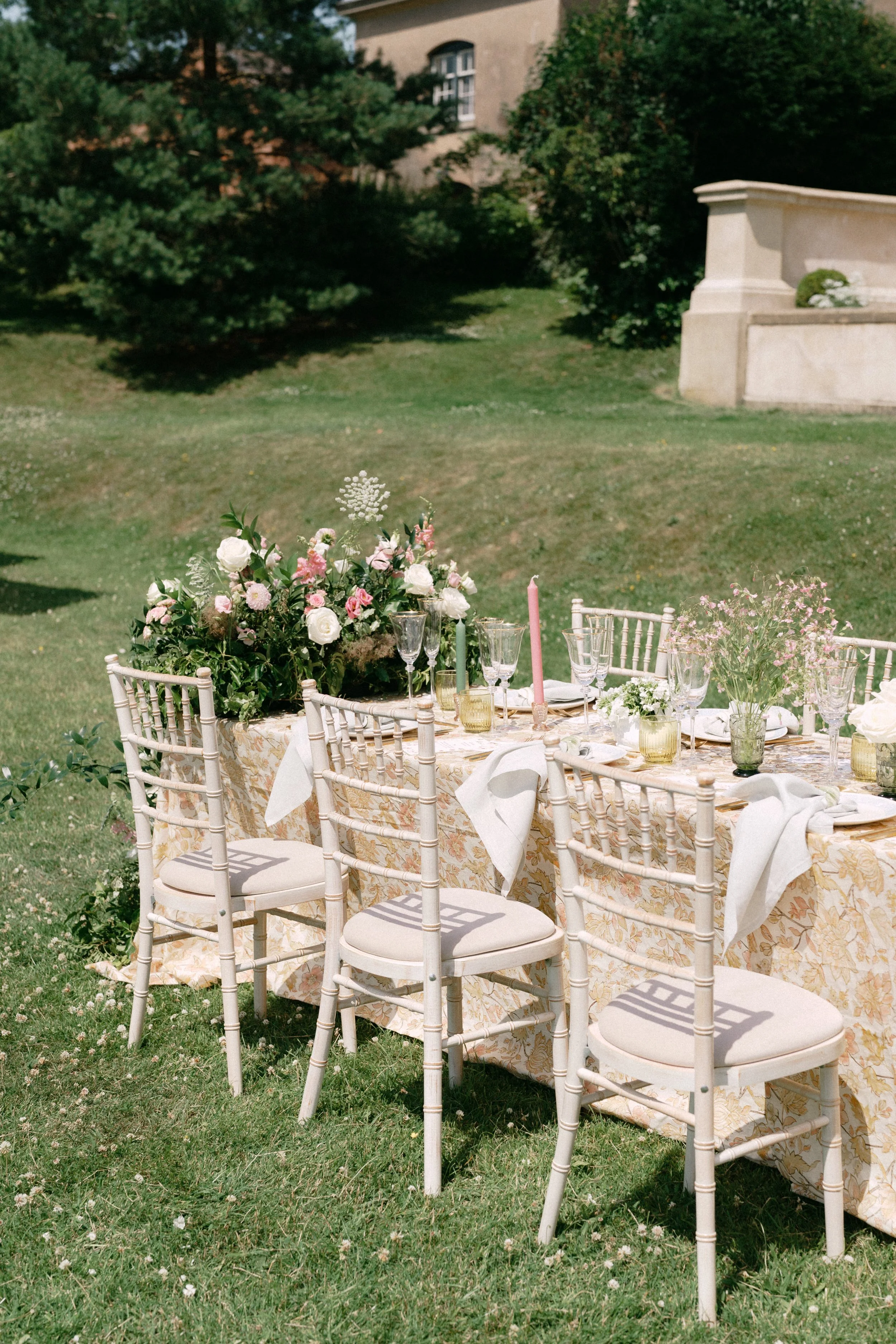 An outdoor wedding or event table decorated with flowers, candles, and glassware, set on grass in a garden with trees and a building in the background.