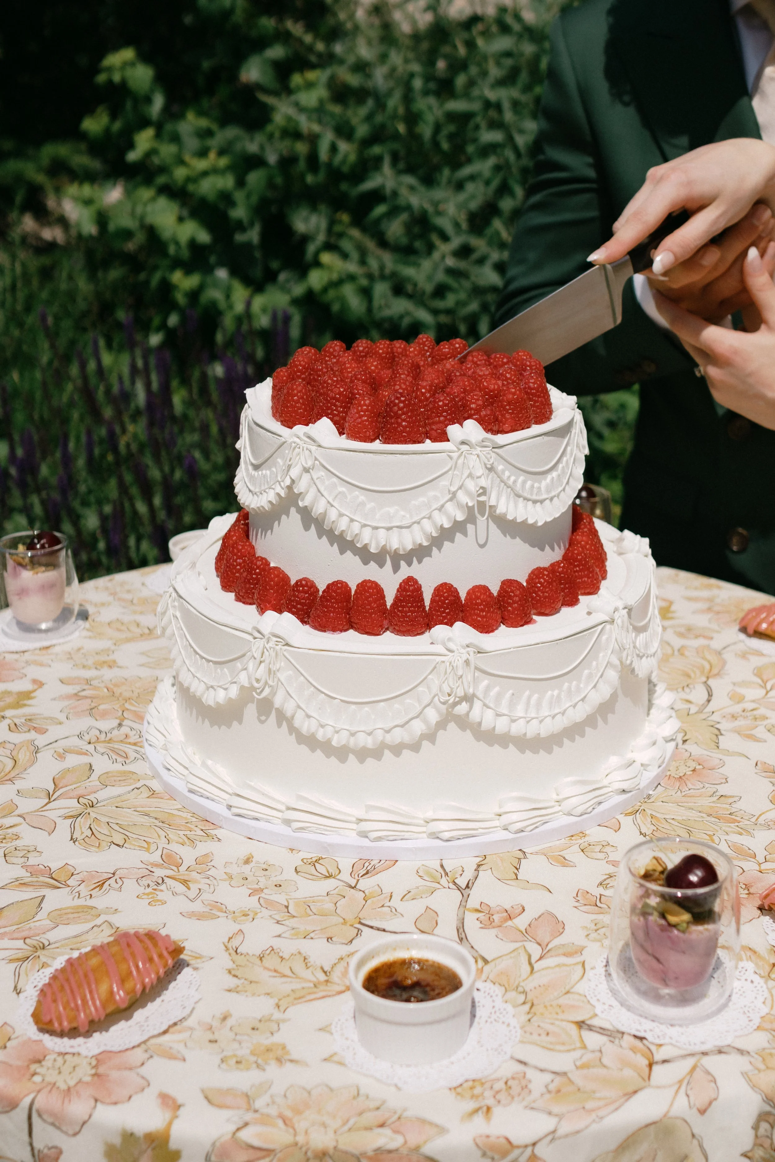 A person in formal attire is slicing a large white cake decorated with red raspberries on top and white icing with ornate piping details. The cake is on a table with a floral tablecloth, surrounded by small desserts and drinks.