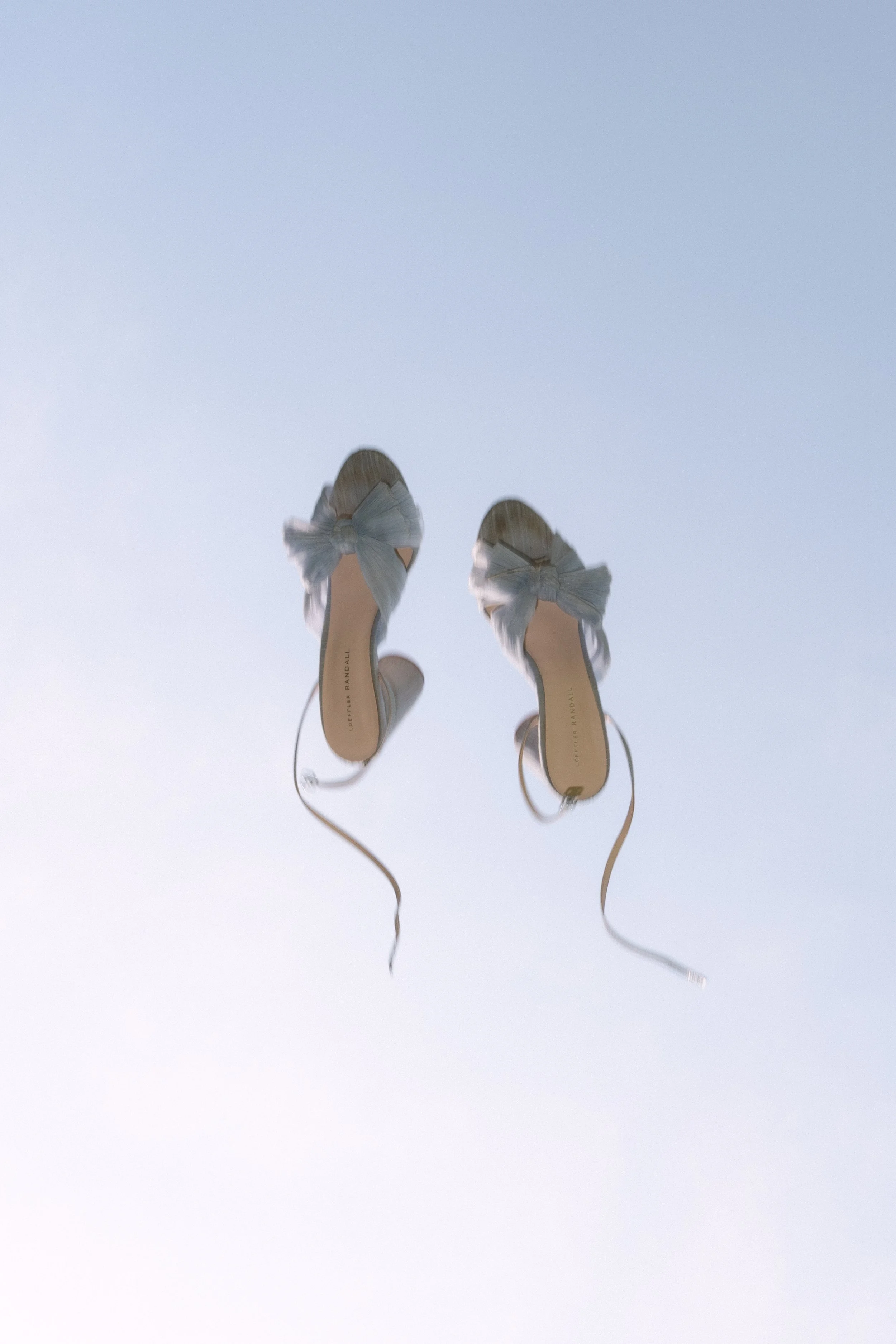 Pair of white high-heeled shoes with bows, photographed from below against a pale blue sky.