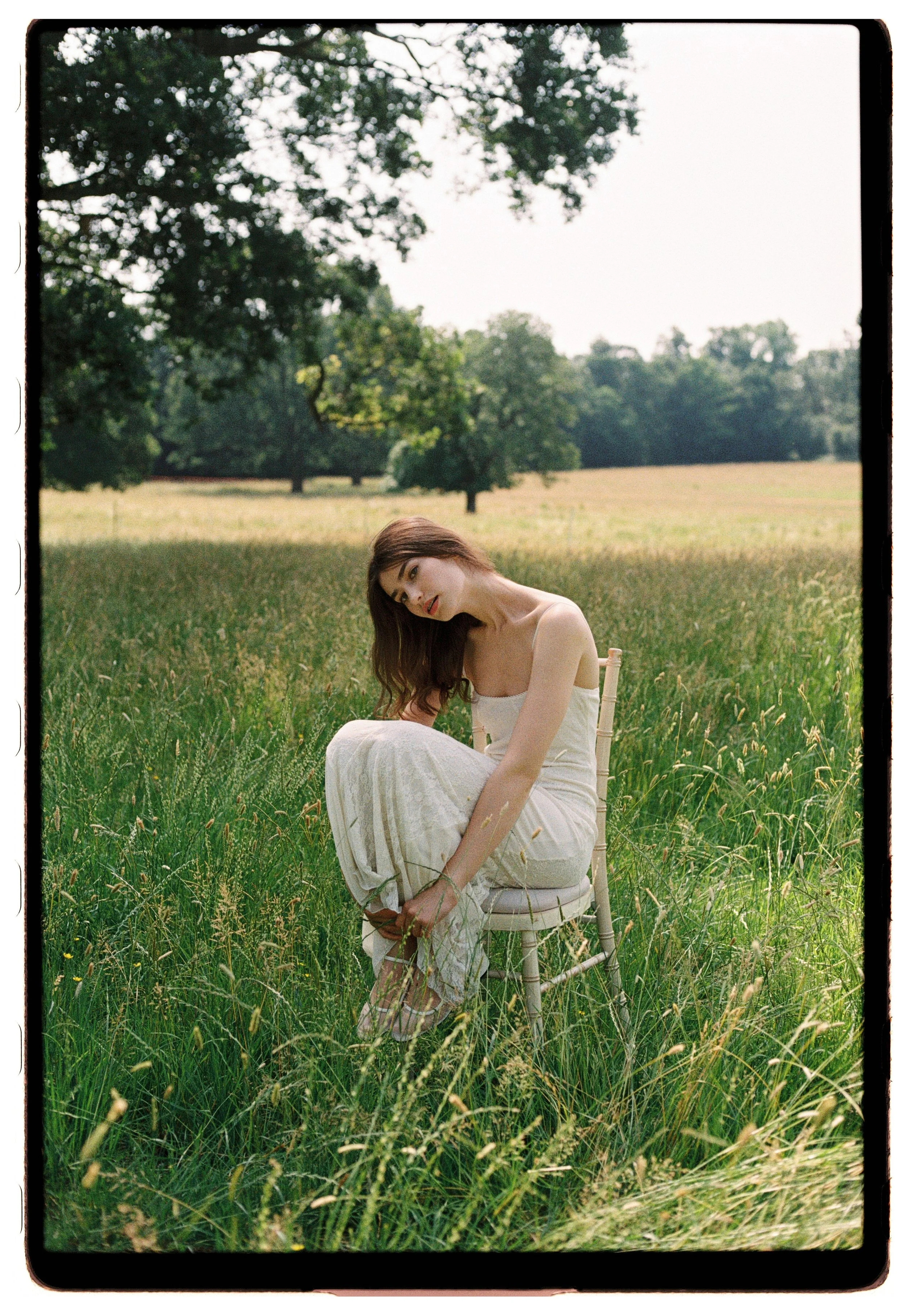 A woman with long, brown hair sitting on a white chair in a grassy field, wearing a white dress, with trees and an open sky in the background.