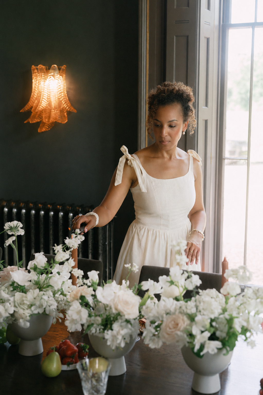A woman in a cream-colored dress arranging white flowers on a dining table, with a dark wall, a window, and a decorative wall light in the background.