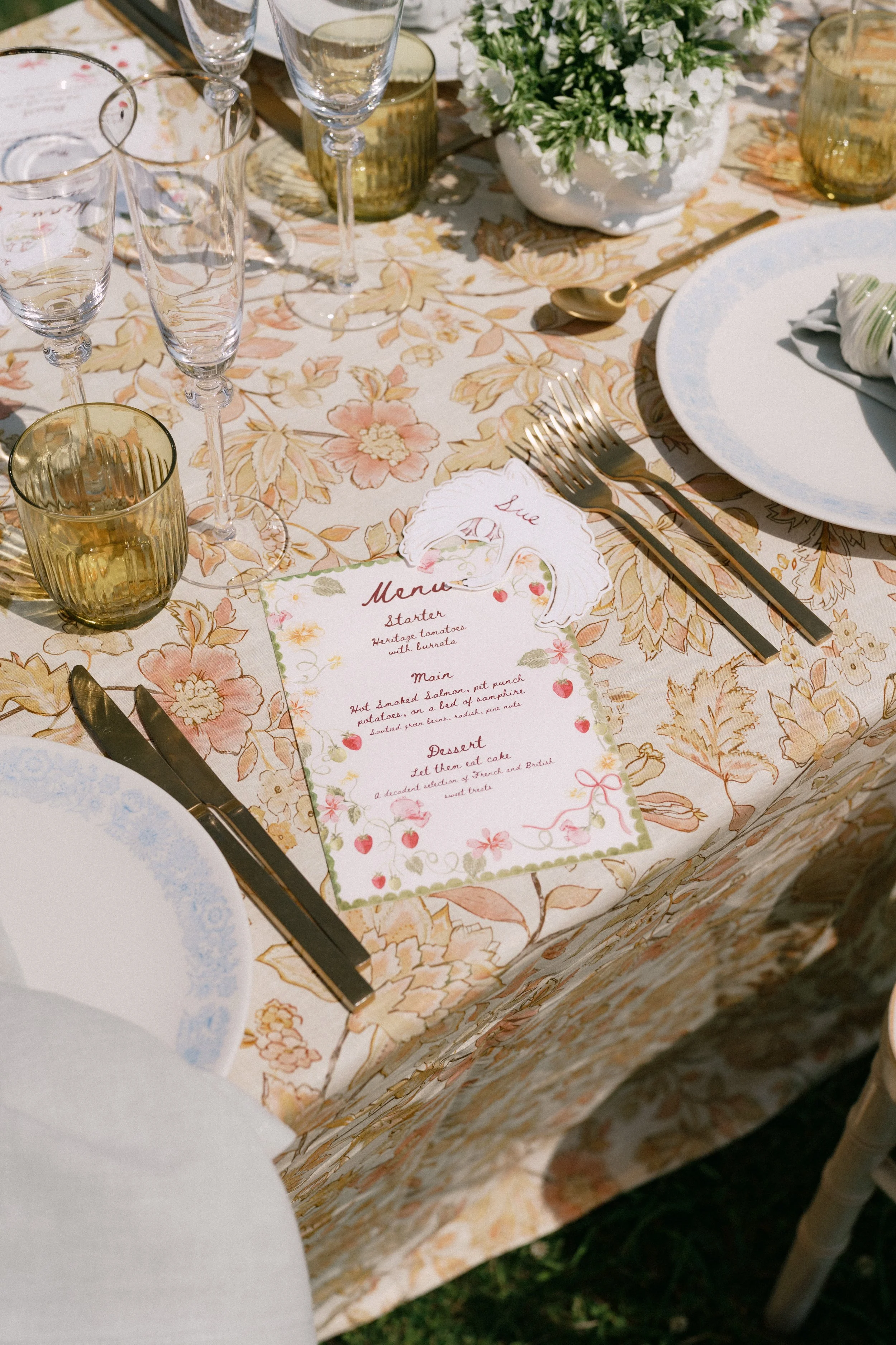 A table set for a meal with floral tablecloth, a white floral arrangement, gold utensils, and a menu with floral accents.