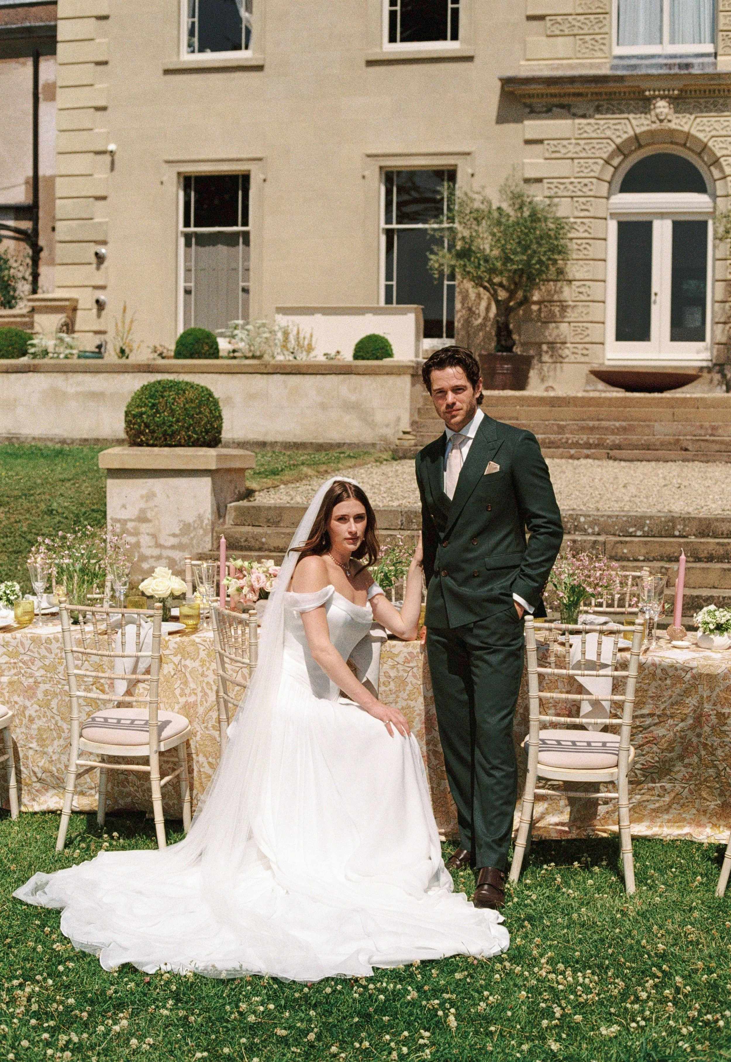 A bride and groom posing outside at a wedding reception in front of a historic building with steps and large windows. The bride is sitting at a decorated table, wearing a white wedding dress with a long train, and the groom is standing next to her in