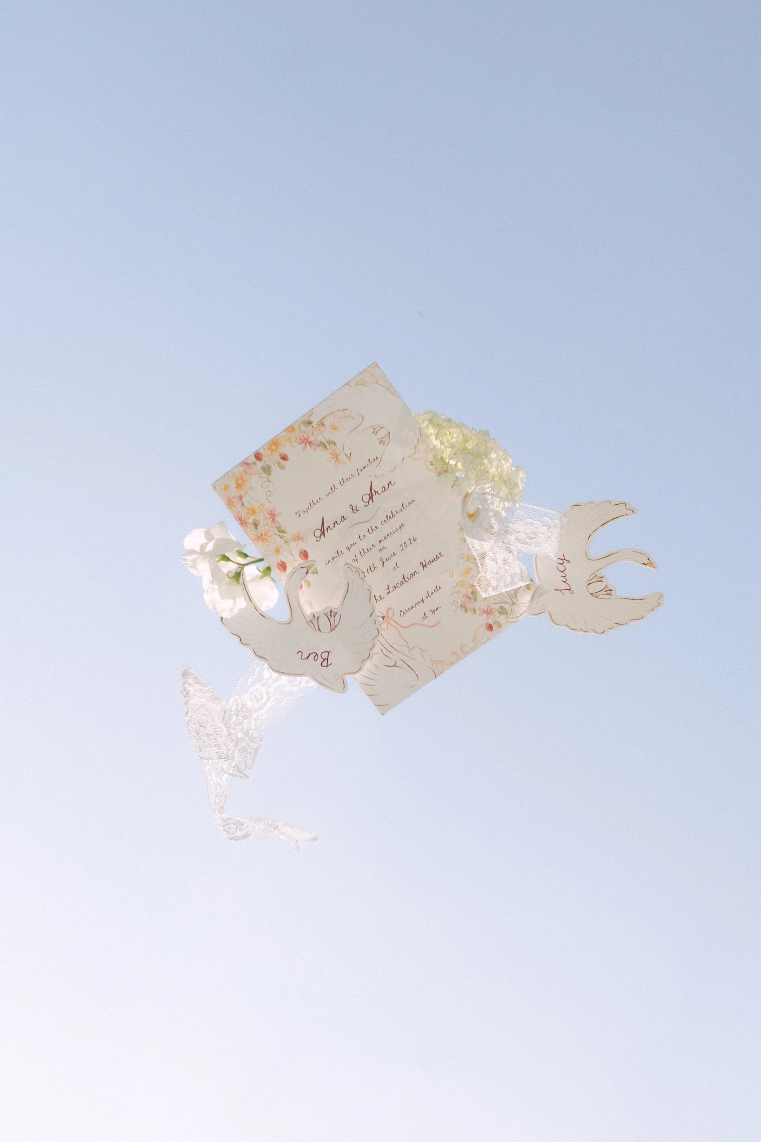 A wedding invitation card with floral design, attached to a white lace and led fairy lights, floating against a clear blue sky.