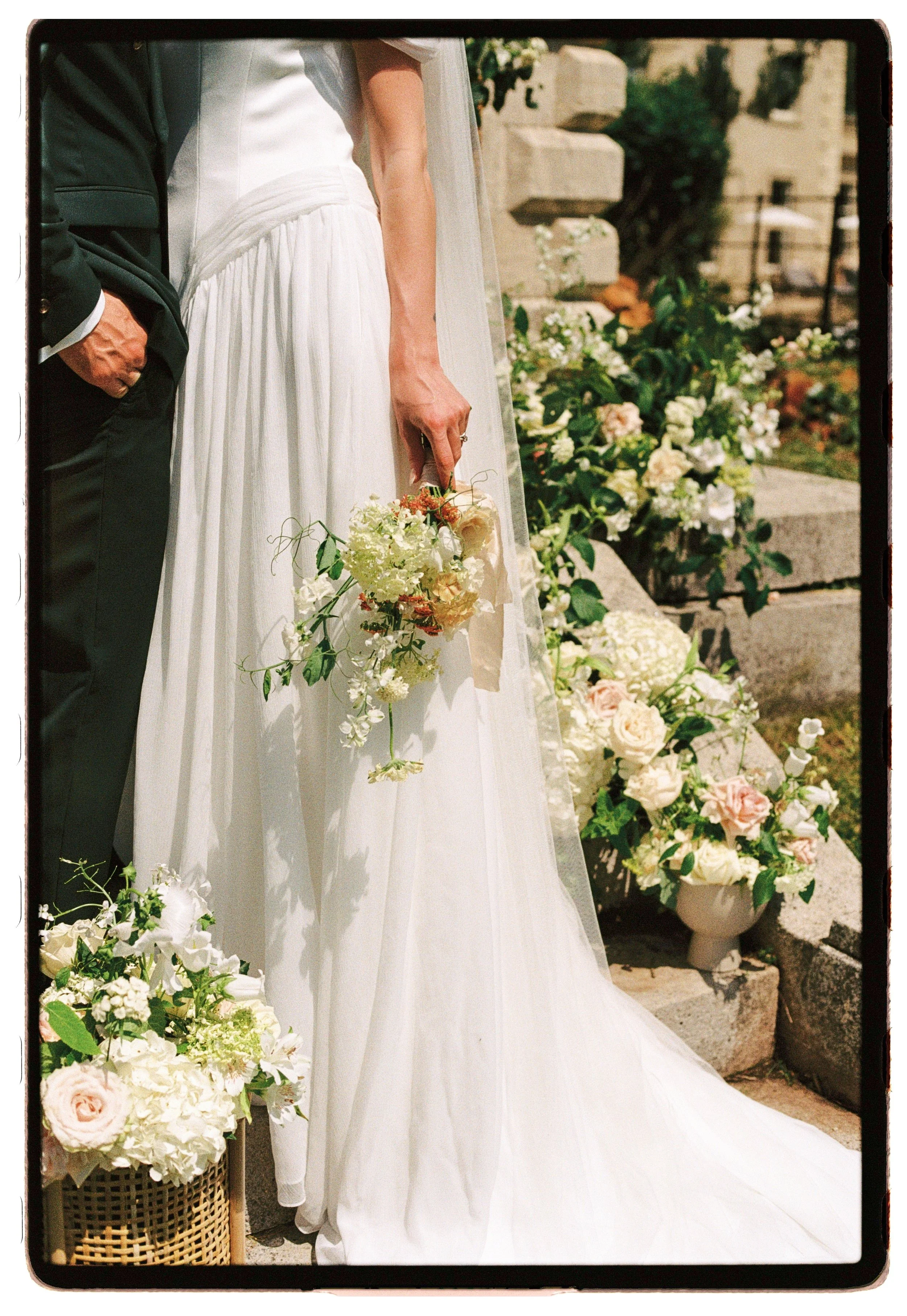 Bride in white wedding dress holding a bouquet standing outside, with flowers and greenery around.