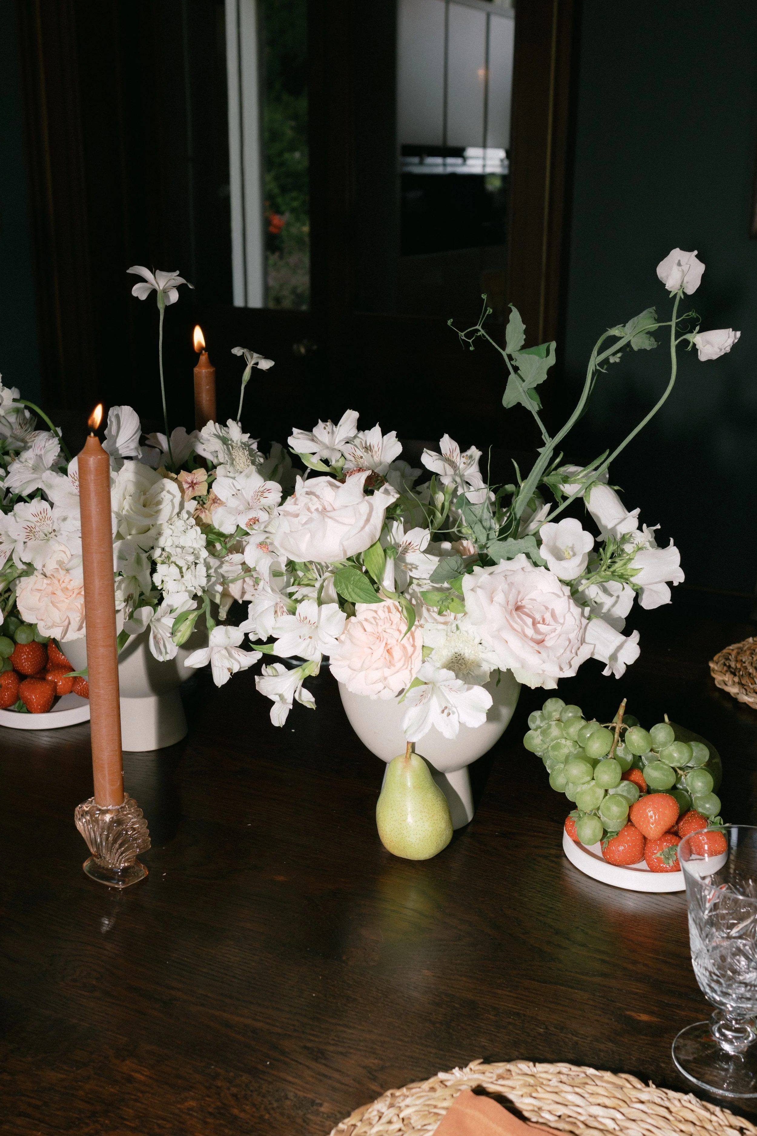 A dining table decorated with white flowers, candles, grapes, strawberries, a pear, and a glass of water, with a dark wooden surface and a mirror in the background.