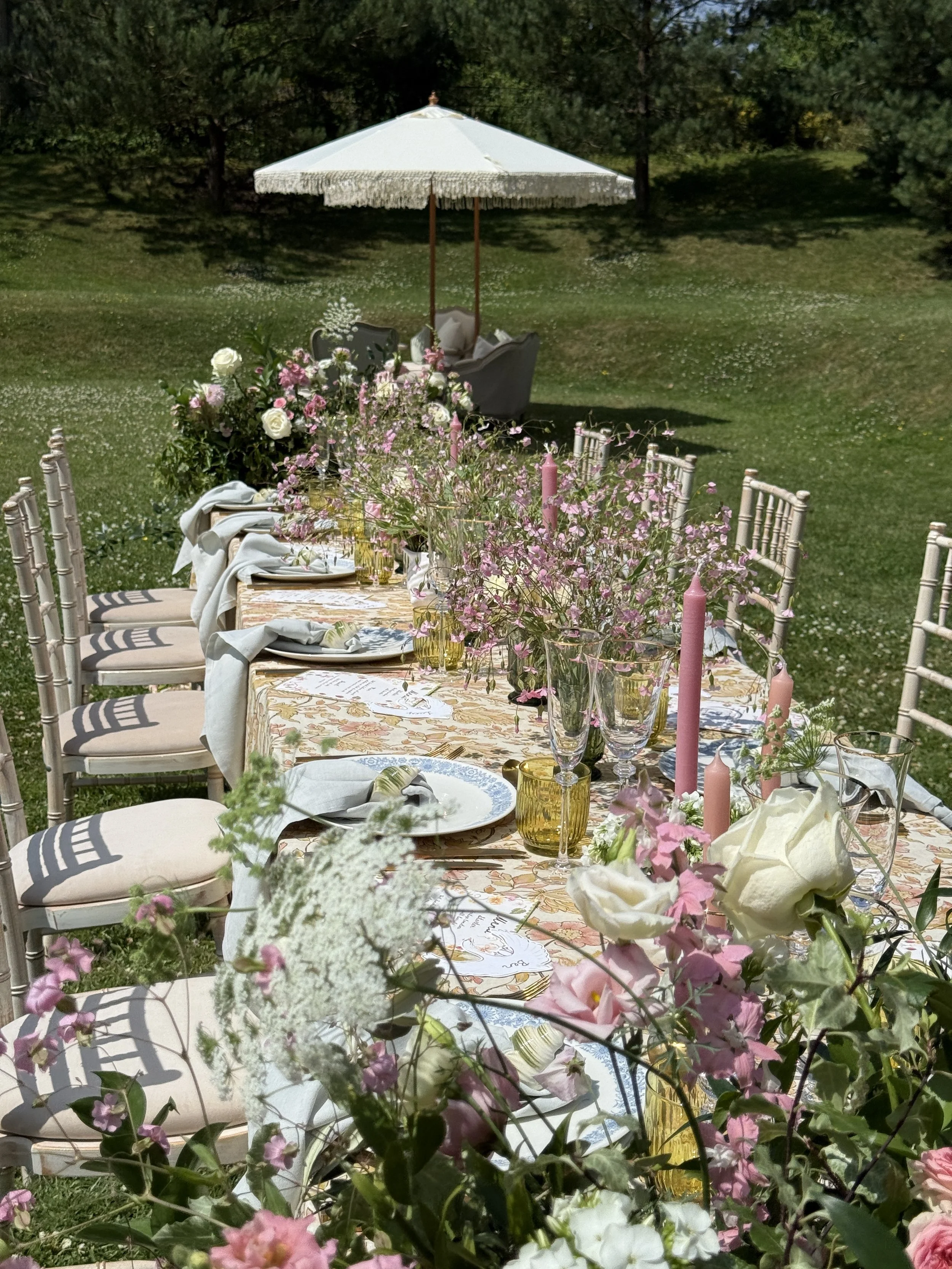 Elegant outdoor dining setup with a long table decorated with flowers, candles, and tableware, surrounded by white chairs, on a lush green lawn with an umbrella in the background.