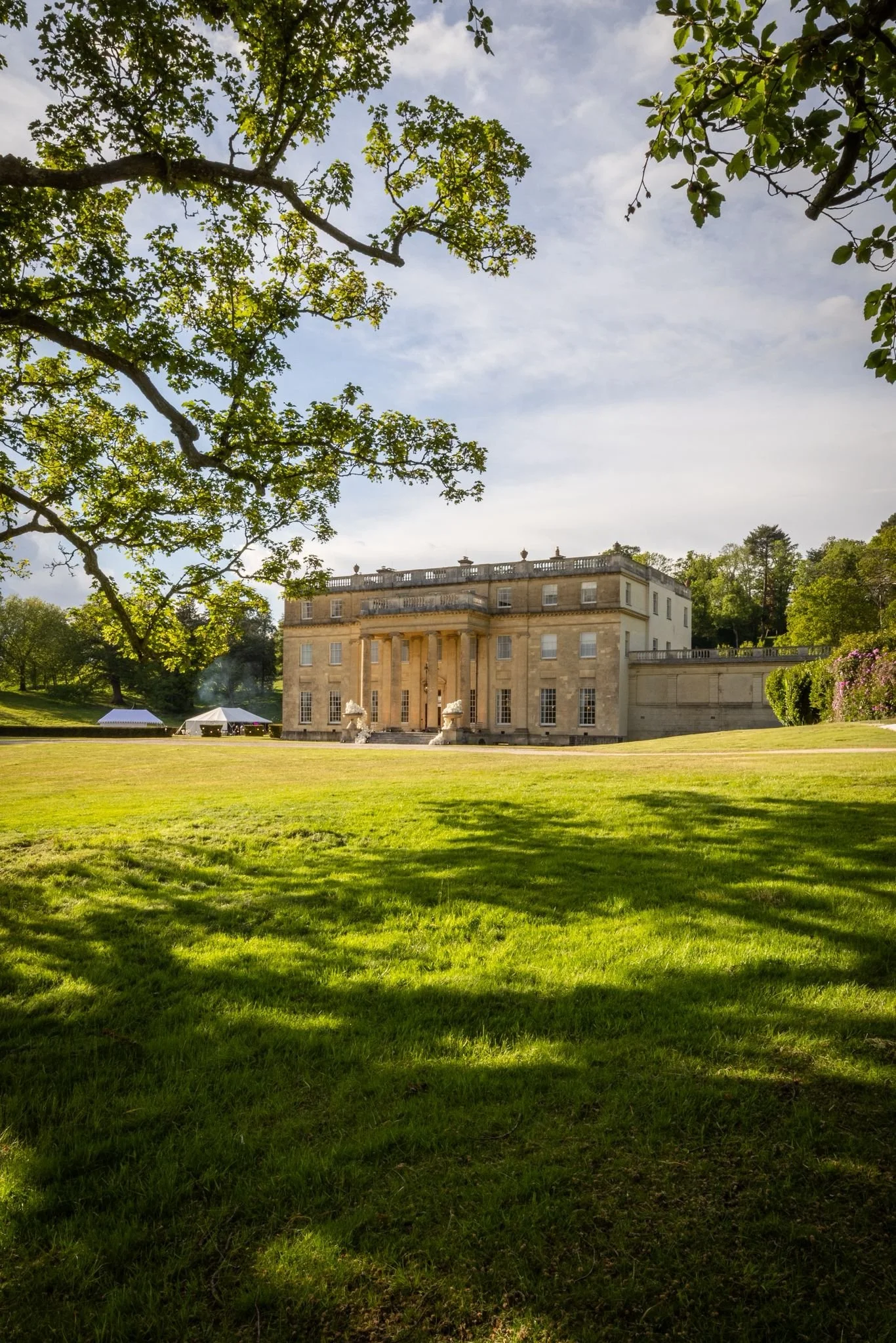 A large neoclassical mansion with columns and balustrades, surrounded by a lush green lawn and trees, under a partly cloudy sky.