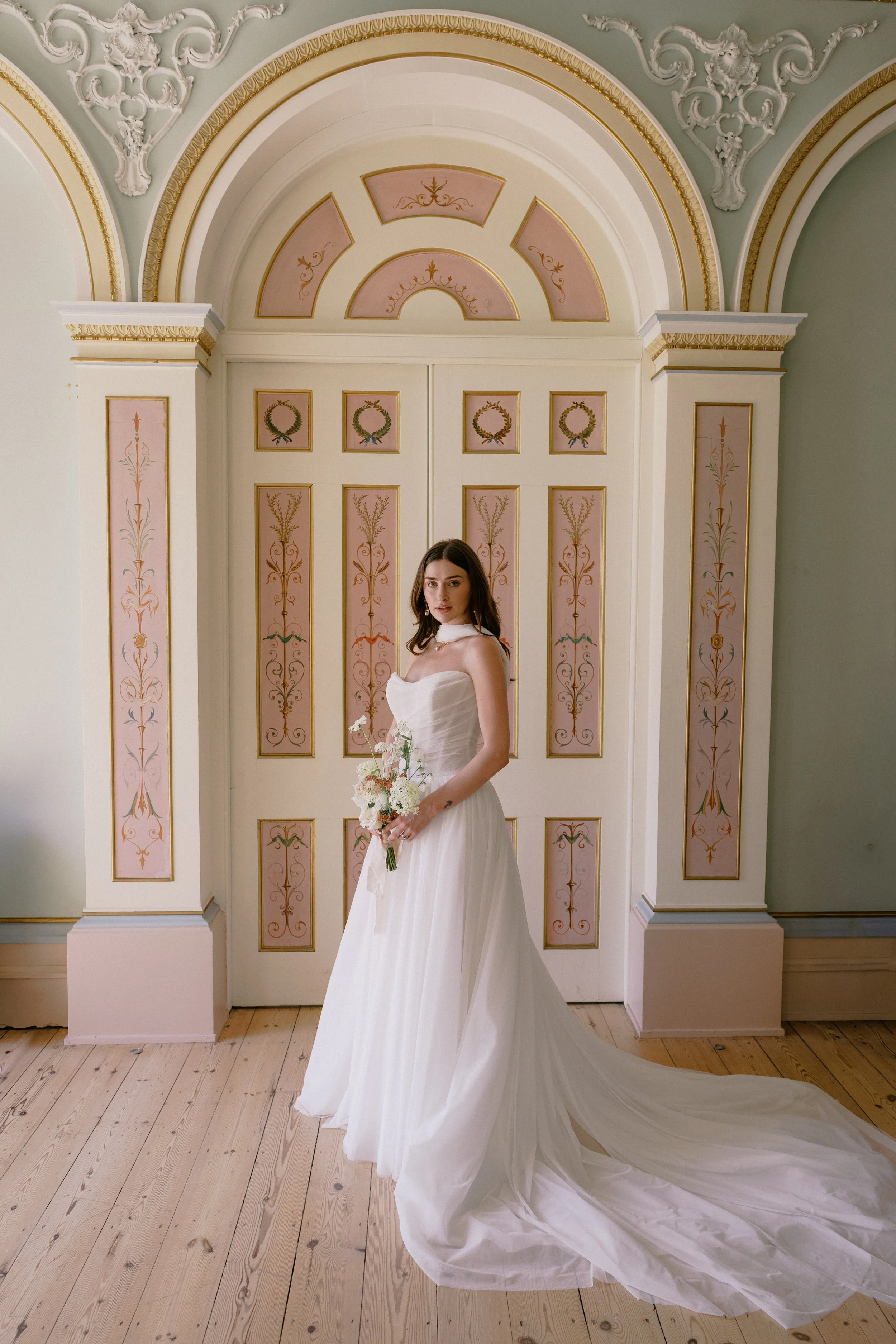 A woman in a wedding dress holding a bouquet stands in front of an ornate, pastel-colored door with gold accents and floral designs, inside a decorated room with intricate plasterwork on the ceiling.