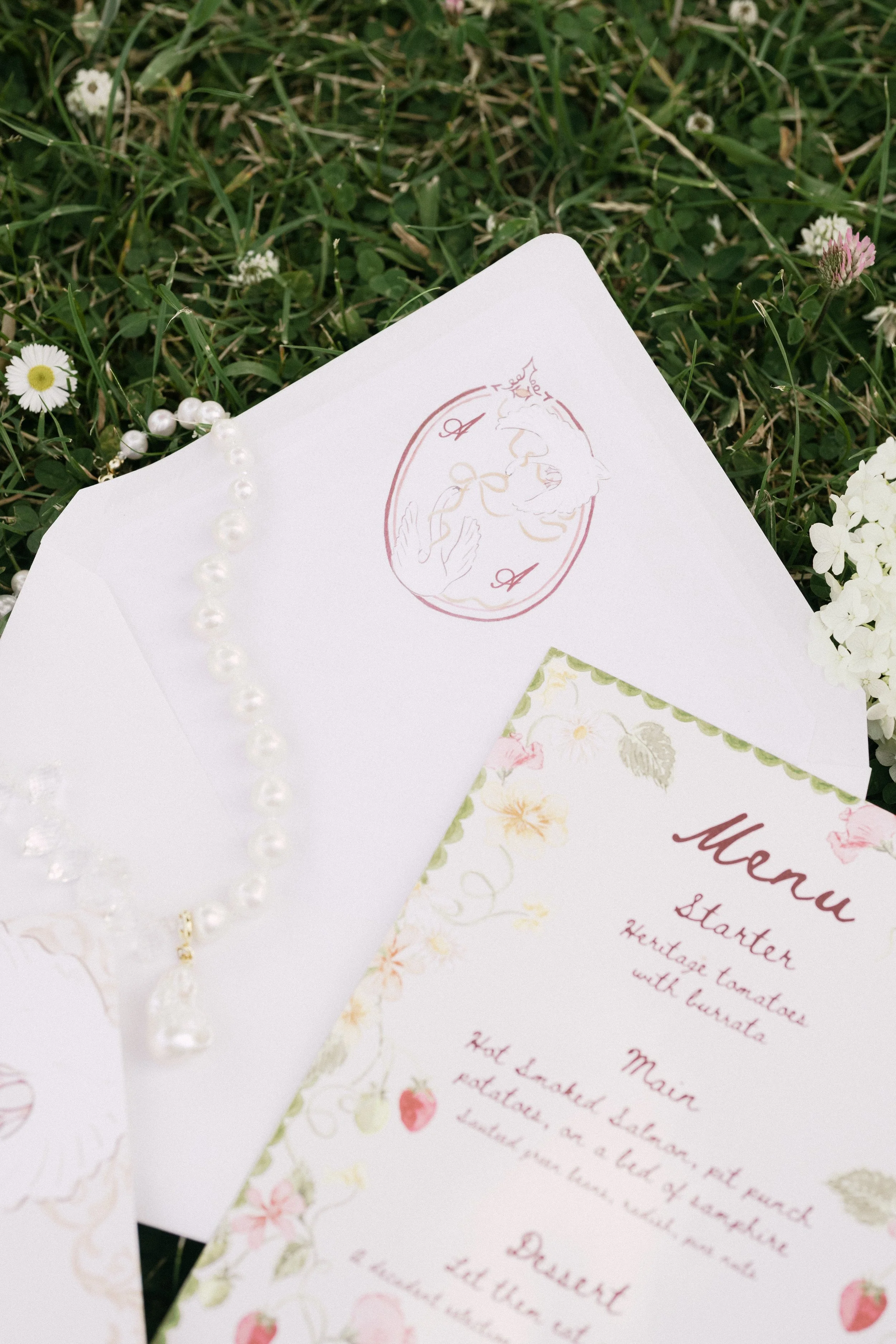 A wedding menu card with floral designs, placed on grass with small flowers, and a pearl necklace nearby.