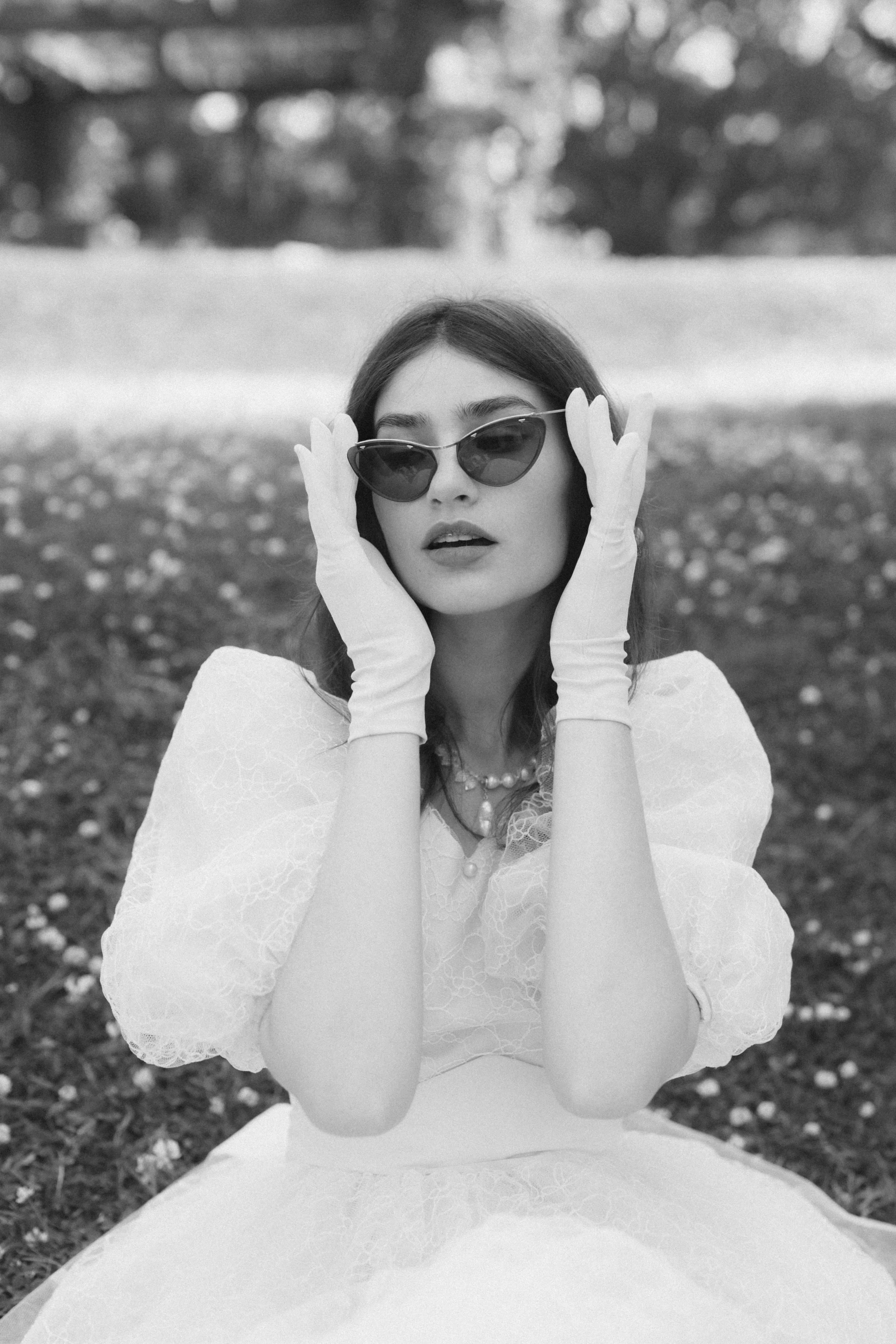 Black and white photo of a woman in a wedding dress, wearing sunglasses, with gloves on her hands, sitting outdoors on the grass with trees in the background.