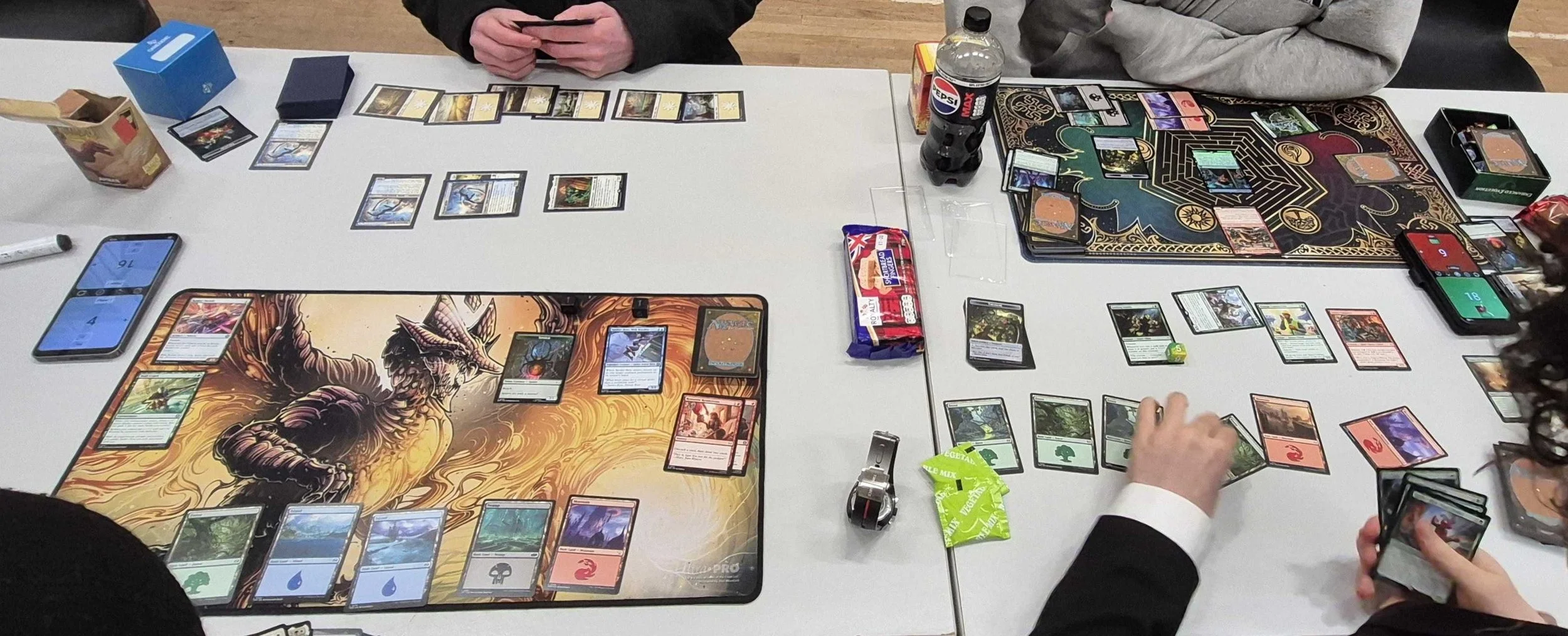 People playing a card game, likely Magic: The Gathering, on a table with various cards, game mats, and drinks, including a bottle of Pepsi.