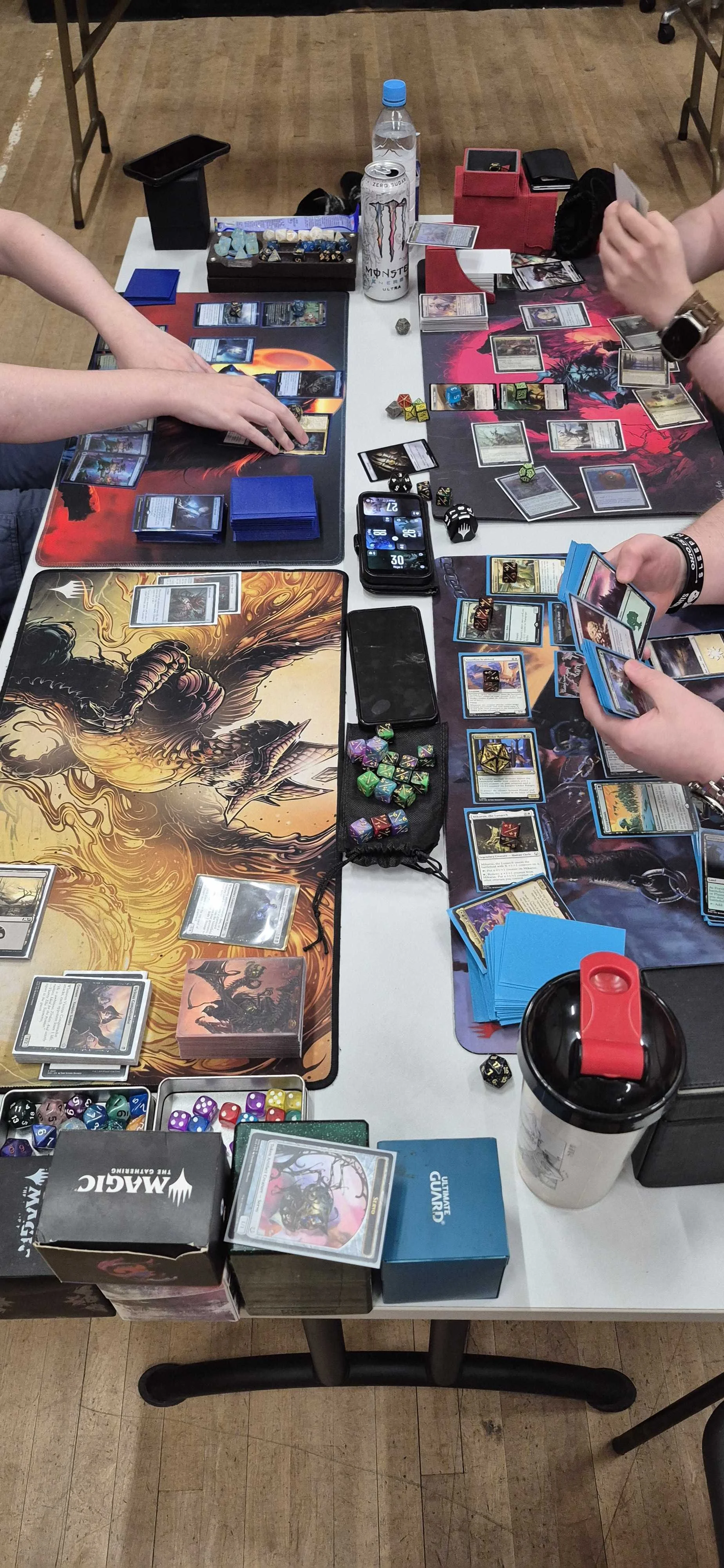 Two players engaged in a card game or trading card game at a table with various game cards, dice, tokens, and accessories. The table has colorful mats featuring dragon artwork, and there are water bottles, a tumbler, and storage boxes around.