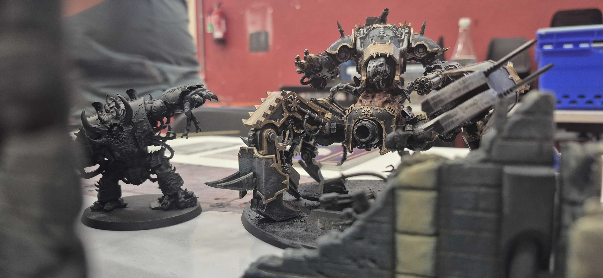 A tabletop scene featuring intricately painted miniatures from a sci-fi or fantasy wargame. Prominent are two figures: a large, alien-like creature with multiple limbs and a heavily armored, mechanical-looking robot with weapons. The background shows workshop or hobby space environment with a red wall, a fire extinguisher, and various supplies.