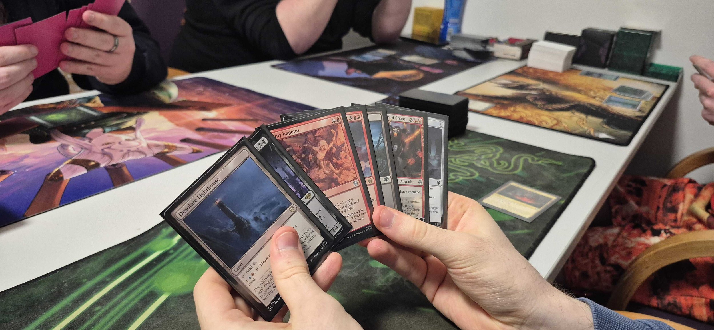 People playing Magic: The Gathering card game, showing hands of cards, game mats, and playing area on the table.