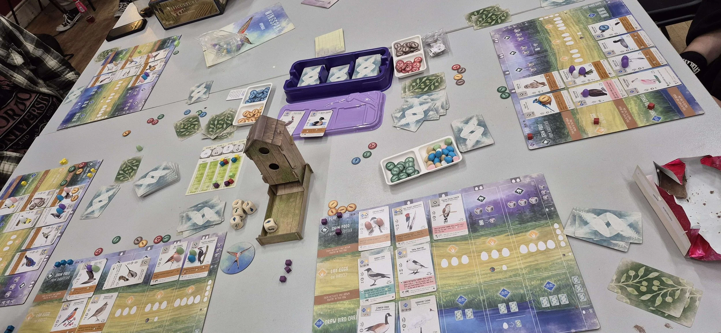 A tabletop board game setup with colorful cards, tokens, and game pieces spread out evenly, with some players' hands visible on each side of the table.