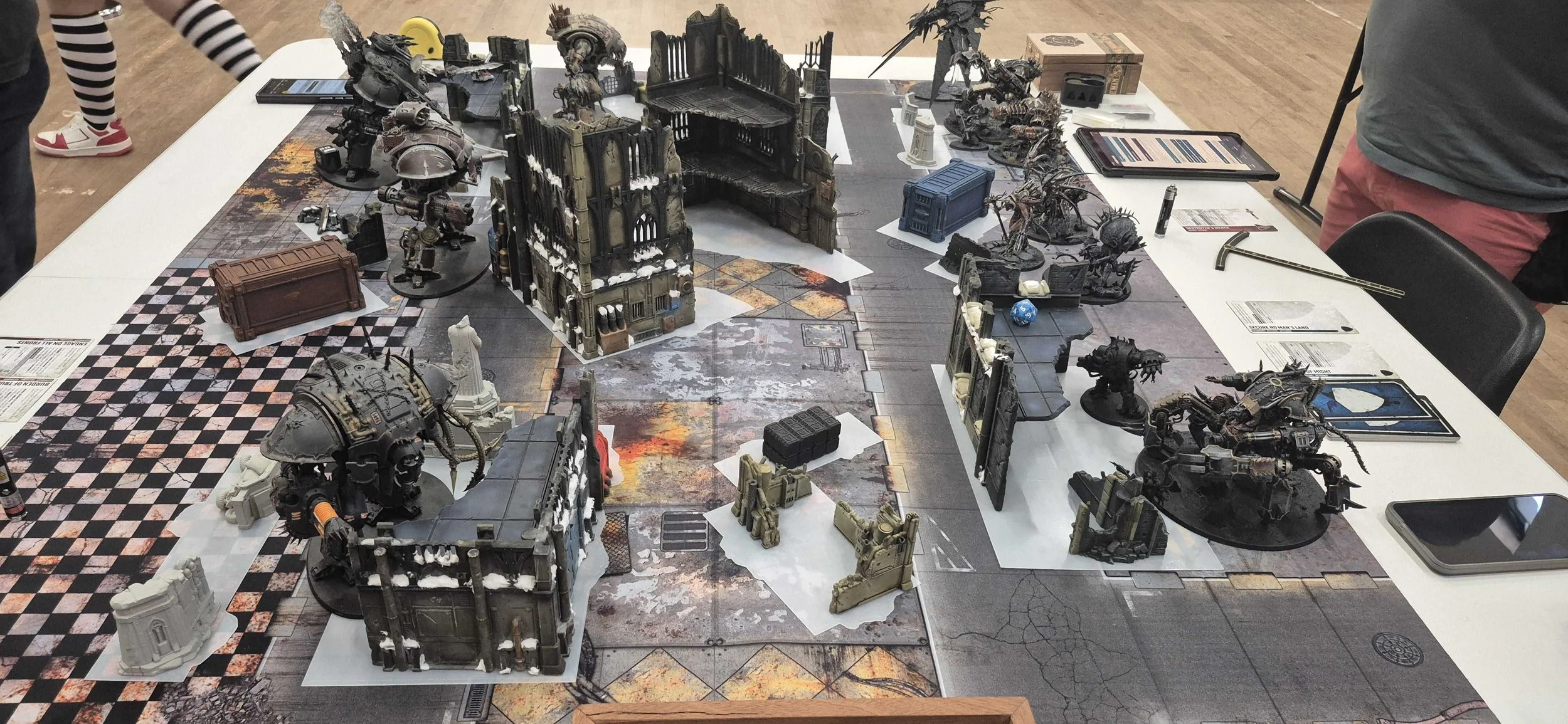 Tabletop gaming setup with detailed miniature buildings, characters, and terrain pieces for a fantasy or sci-fi game, with game cards, dice, and tablets nearby.