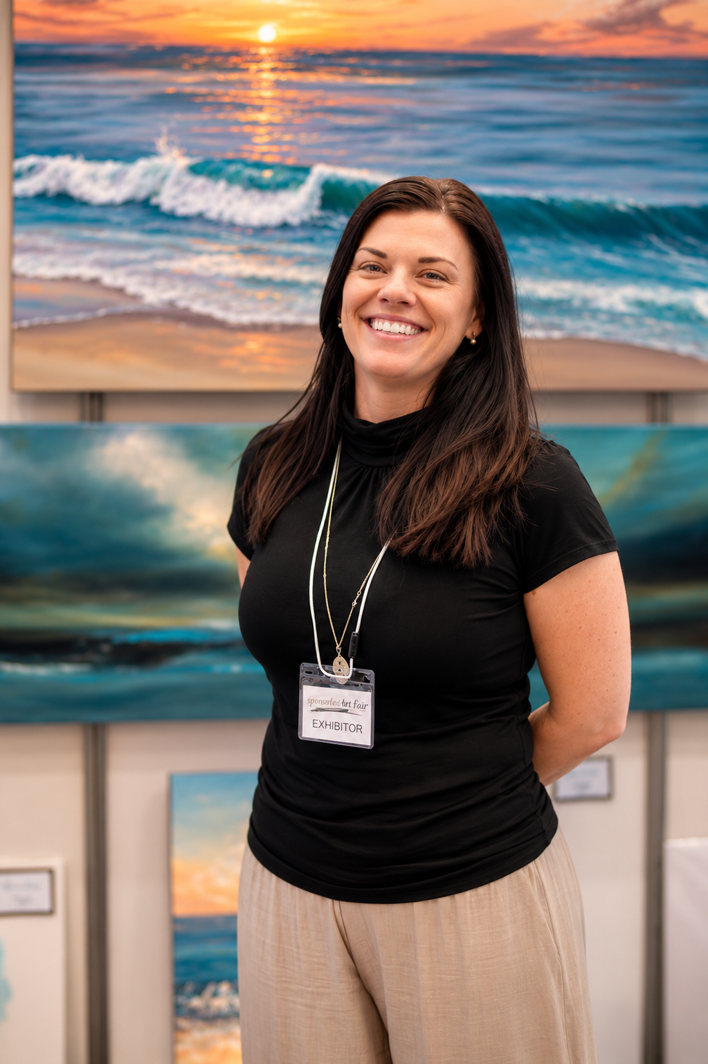 A smiling woman standing in front of paintings of ocean scenes at an art fair. She is wearing a black top, beige pants, and an exhibitor badge.