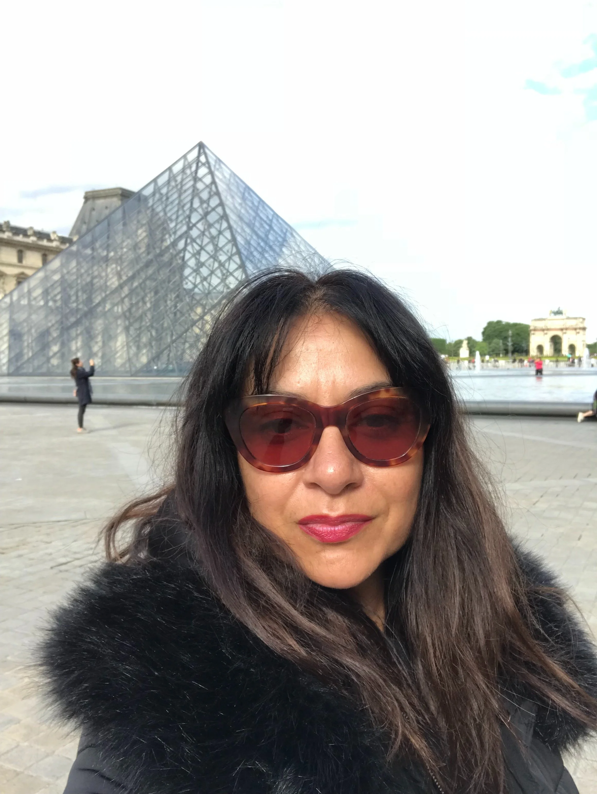 A woman wearing sunglasses and a black fur coat taking a selfie in front of the Louvre Pyramid with the Louvre Museum in the background.