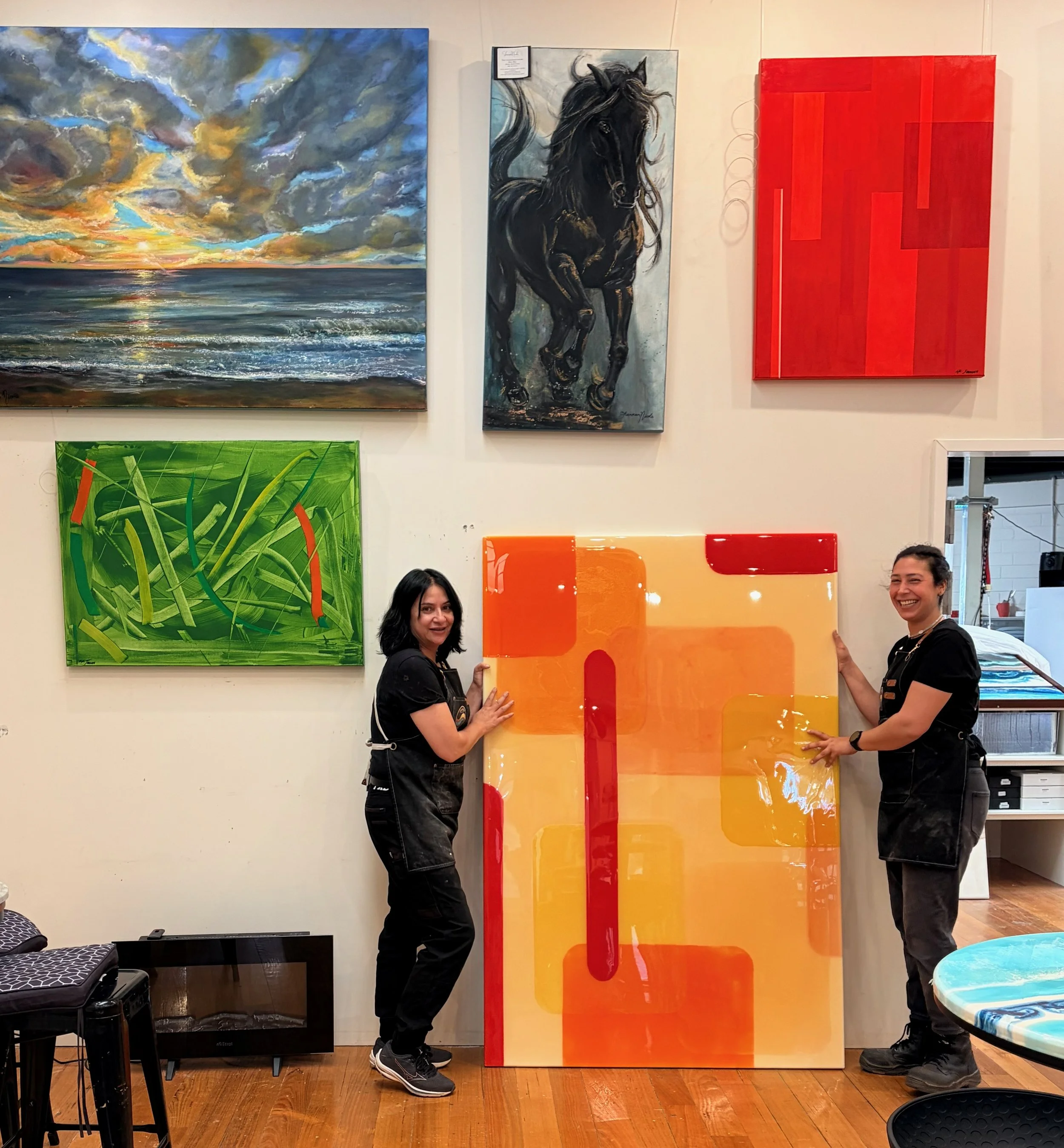 Two women are standing inside an art gallery, each holding a large piece of artwork. The woman on the left has black hair, is wearing black pants and shoes, and stands next to a colorful abstract painting. The woman on the right has dark hair, is wea
