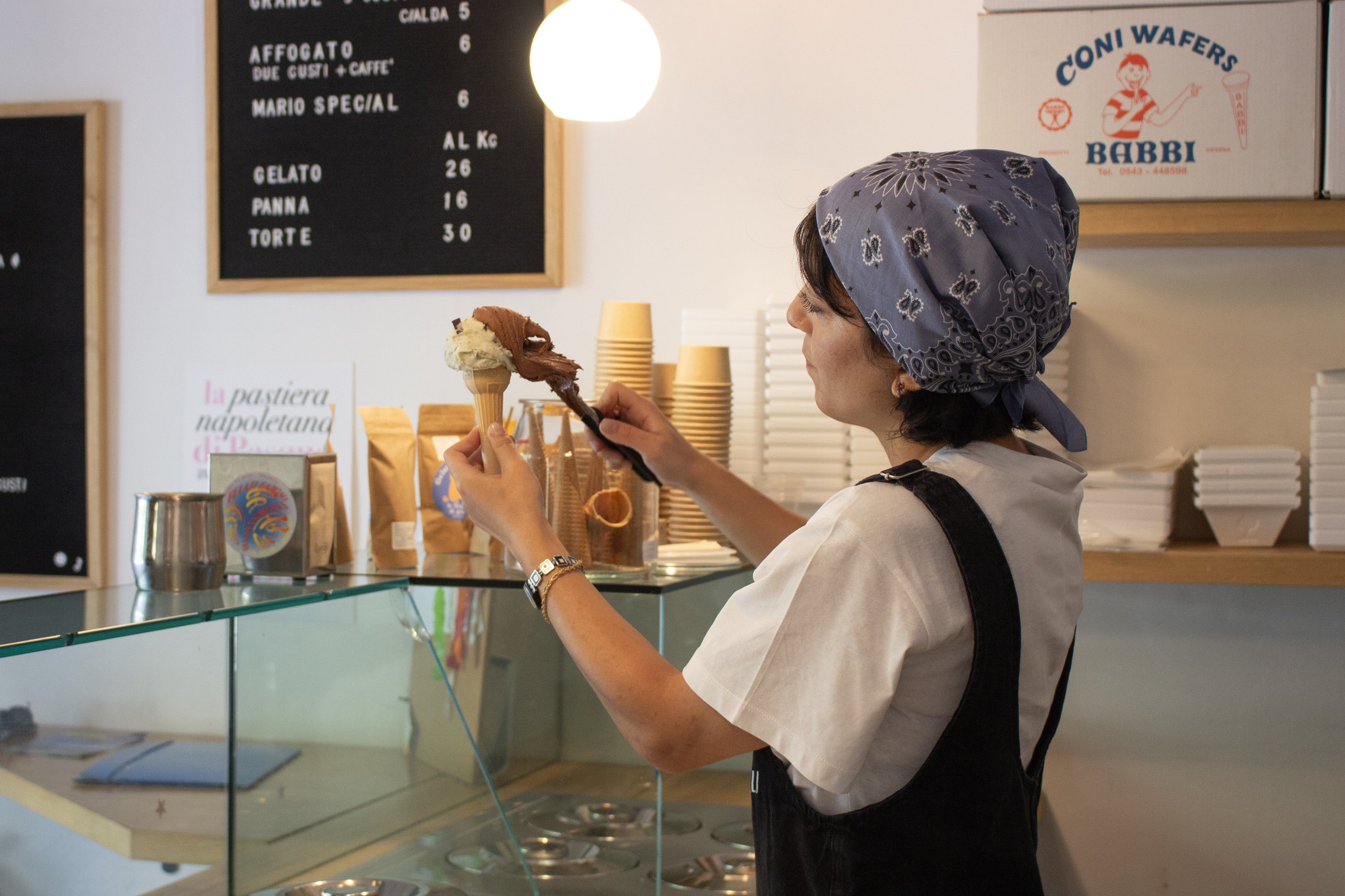 Una donna con un foulard azzurro che indossa un grembiule nero, mentre tiene in mano un bicchiere di gelato nel negozio di coni wafers Barbi. Sullo sfondo ci sono un quadro con il menù, alcune scatole e recipienti, e un cartello pubblicitario con il disegno di una bambina e la scritta 'Coni Wafers Babbi'.