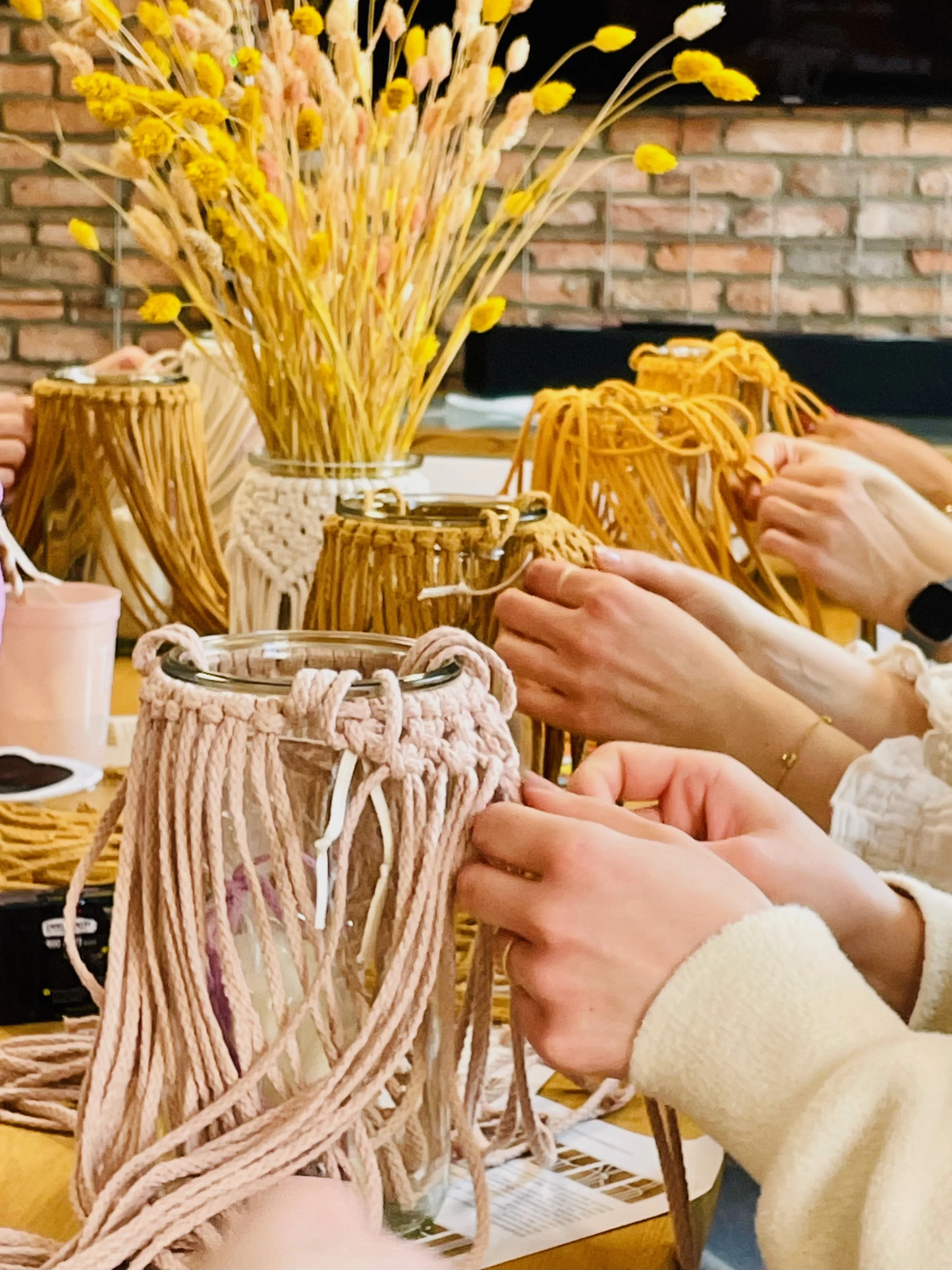 People making a craft, weaving or braiding yarn or string at a table with a bouquet of dried yellow and pink flowers in a jar and a brick wall in the background.