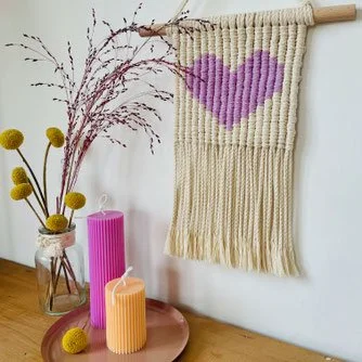 Decorative wall hanging with a purple heart on a beige background, hanging above a wooden table with candles and a vase of flowers.