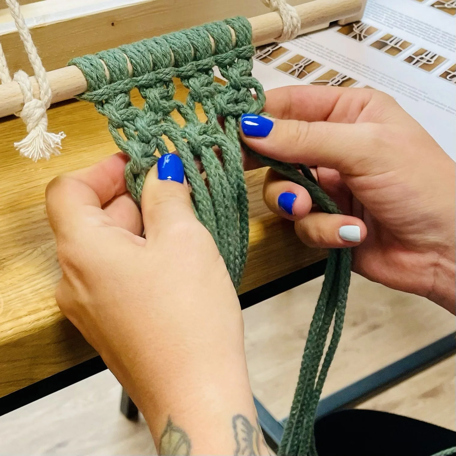 Person with blue and white painted nails knitting green yarn on a wooden loom from a pattern or instruction booklet.