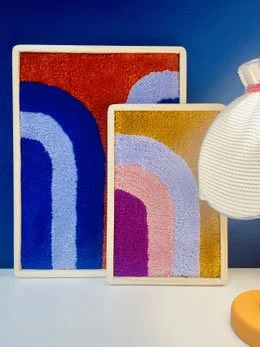 Two abstract colorful paintings with rainbow-like patterns, one larger with red, blue, and white colors, and one smaller with purple, pink, and yellow hues, placed on a white surface with a modern white textured vase to the side against a blue background.