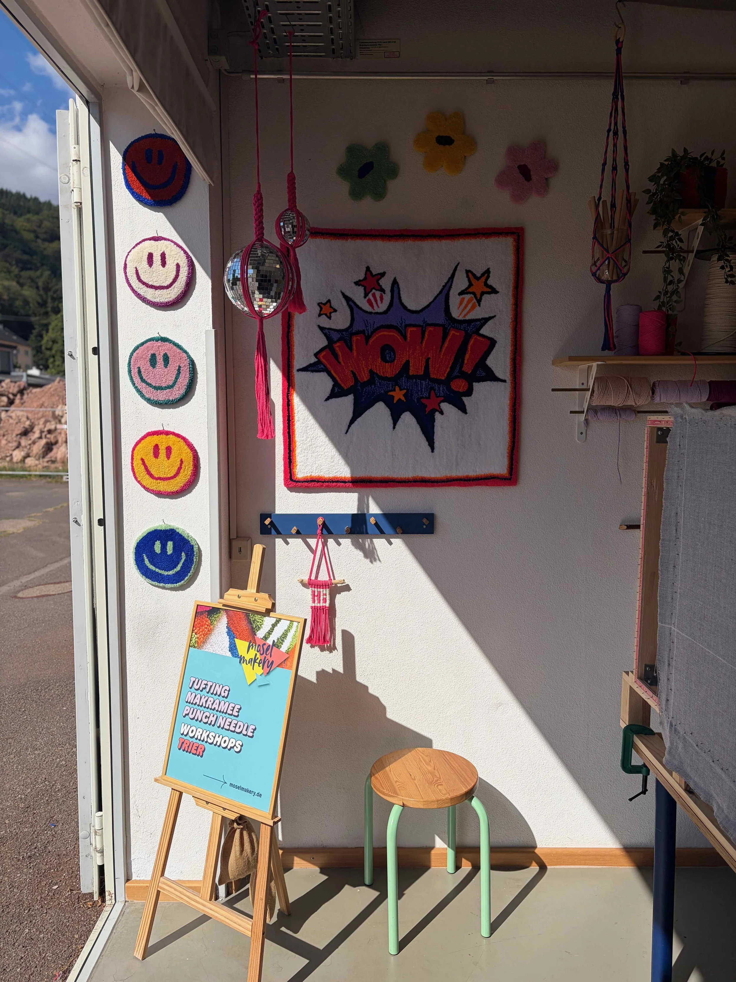 Interior of a craft shop with colorful smiley face wall decorations, a sign with a list of craft activities, a small wooden stool with a round seat, a chalkboard, and crafting supplies on shelves. Natural light coming through an open door.