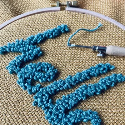 Close-up of a blue knitted or crocheted piece in an embroidery hoop on a textured fabric background, with a crochet tool nearby.