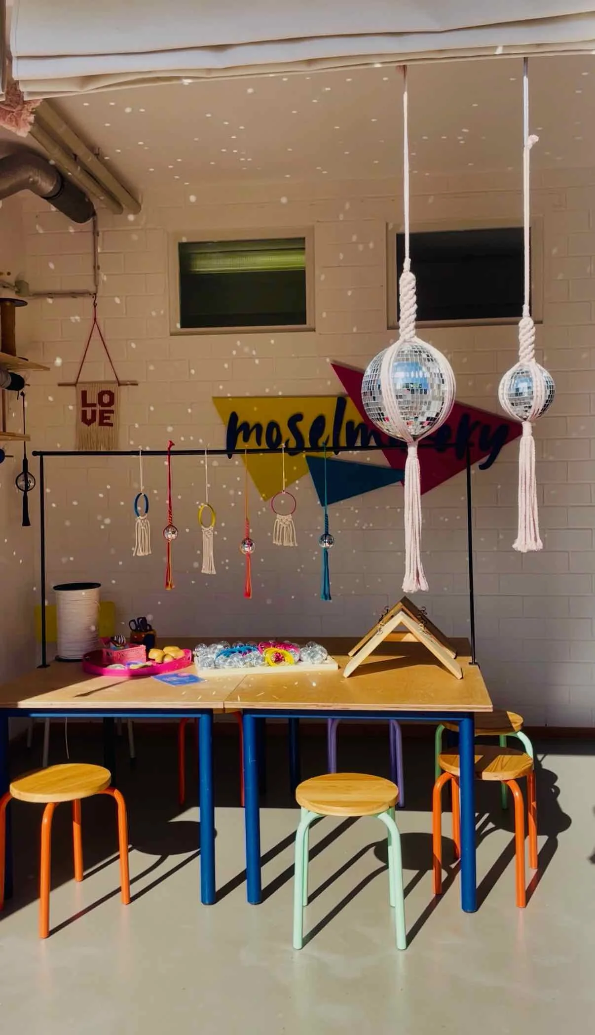 Children's craft display with hanging dreamcatchers, mirror balls, and a sign that says 'moslemery.' There are colorful stools around a wooden table with various craft supplies on it, in a classroom with beige brick walls and small windows.