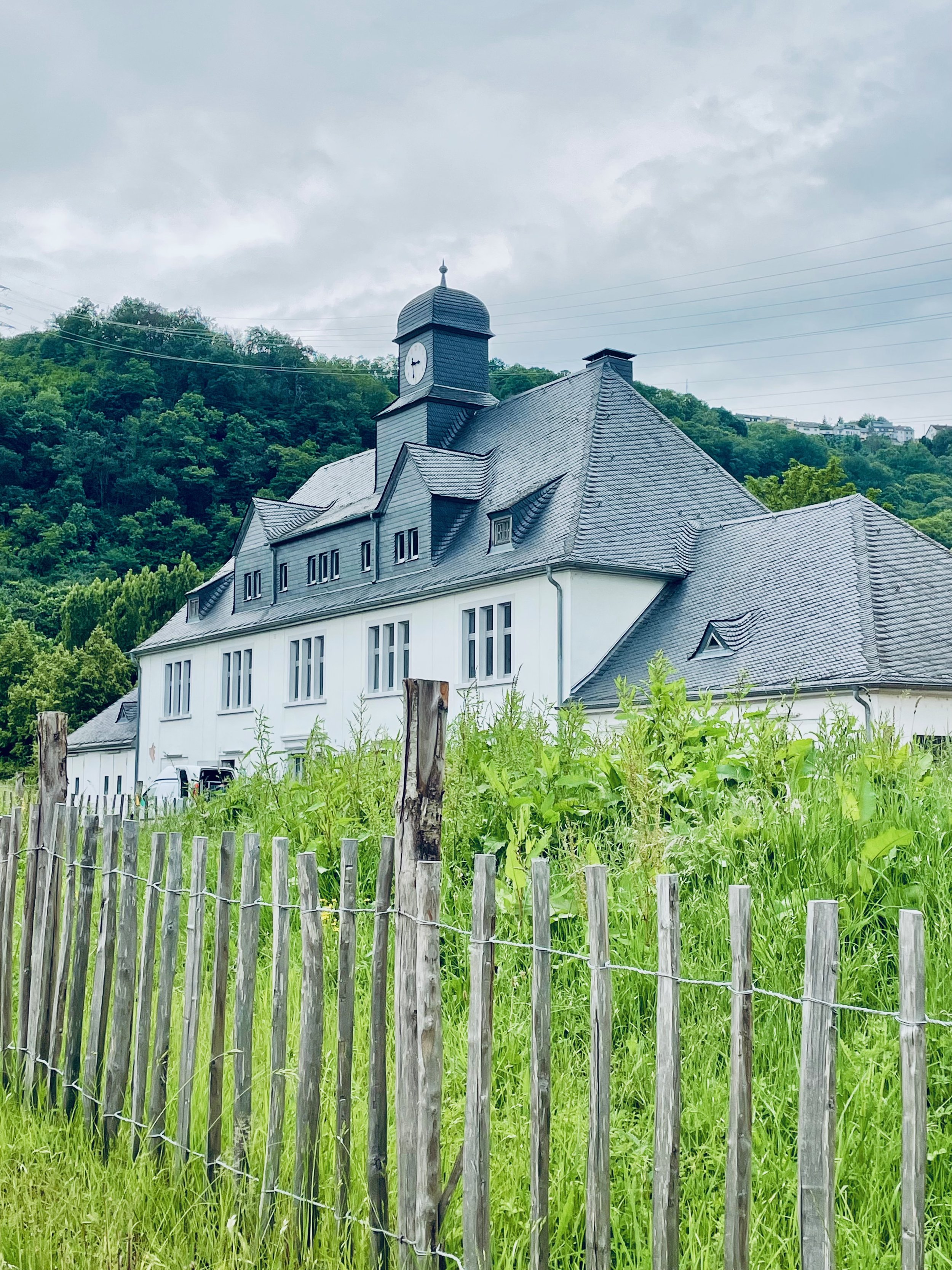 A white building with a slate roof and a clock tower is behind a wooden fence with green grass and trees in the background under a cloudy sky.