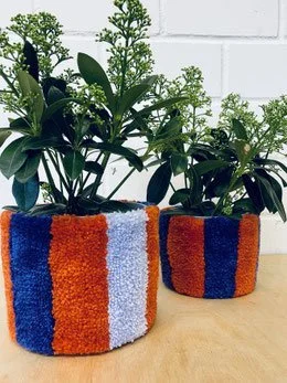 Two small potted plants in colorful knitted or fabric pots with orange, white, and blue stripes on a wooden surface with a white brick wall in the background.