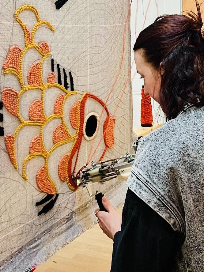 A woman working on a textured, colorful fish tapestry with a hot glue gun. The fish is made of yarn and beads, featuring orange, yellow, and black colors.