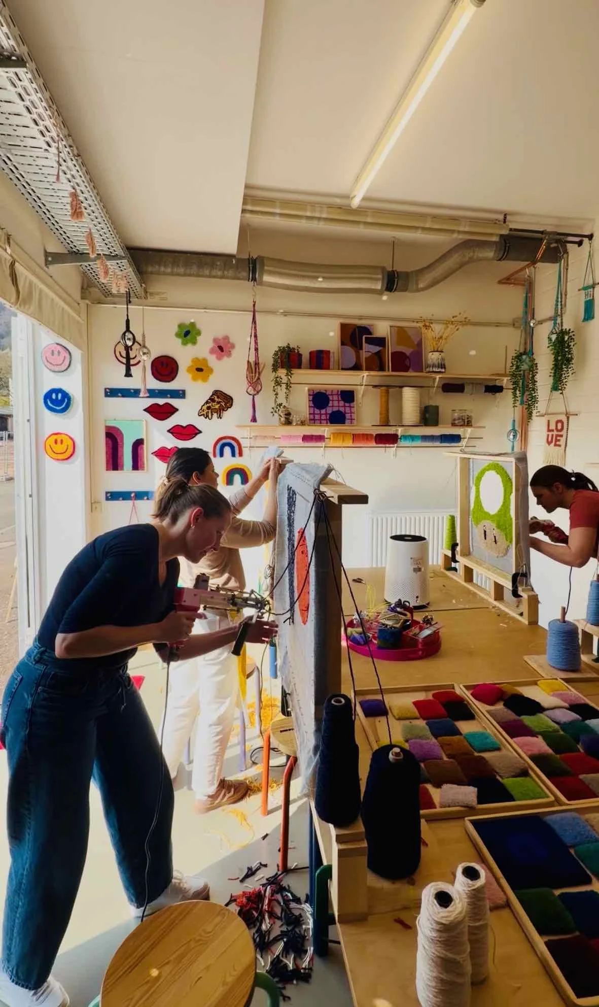 People working on a craft project in a colorful, decorated workshop. Shelves with yarn, fabric, and art supplies. Wall decorations include smiley faces, lips, and rainbows.