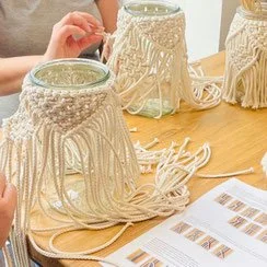 Person making macrame plant hangers with beige cords on a wooden table, alongside printed instructions.