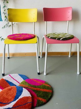 Two colorful chairs, one yellow and one pink, with round cushions on their seats, positioned against a wall near a wooden floor and painted wall. Two patterned round rugs are on the floor in front of the chairs.