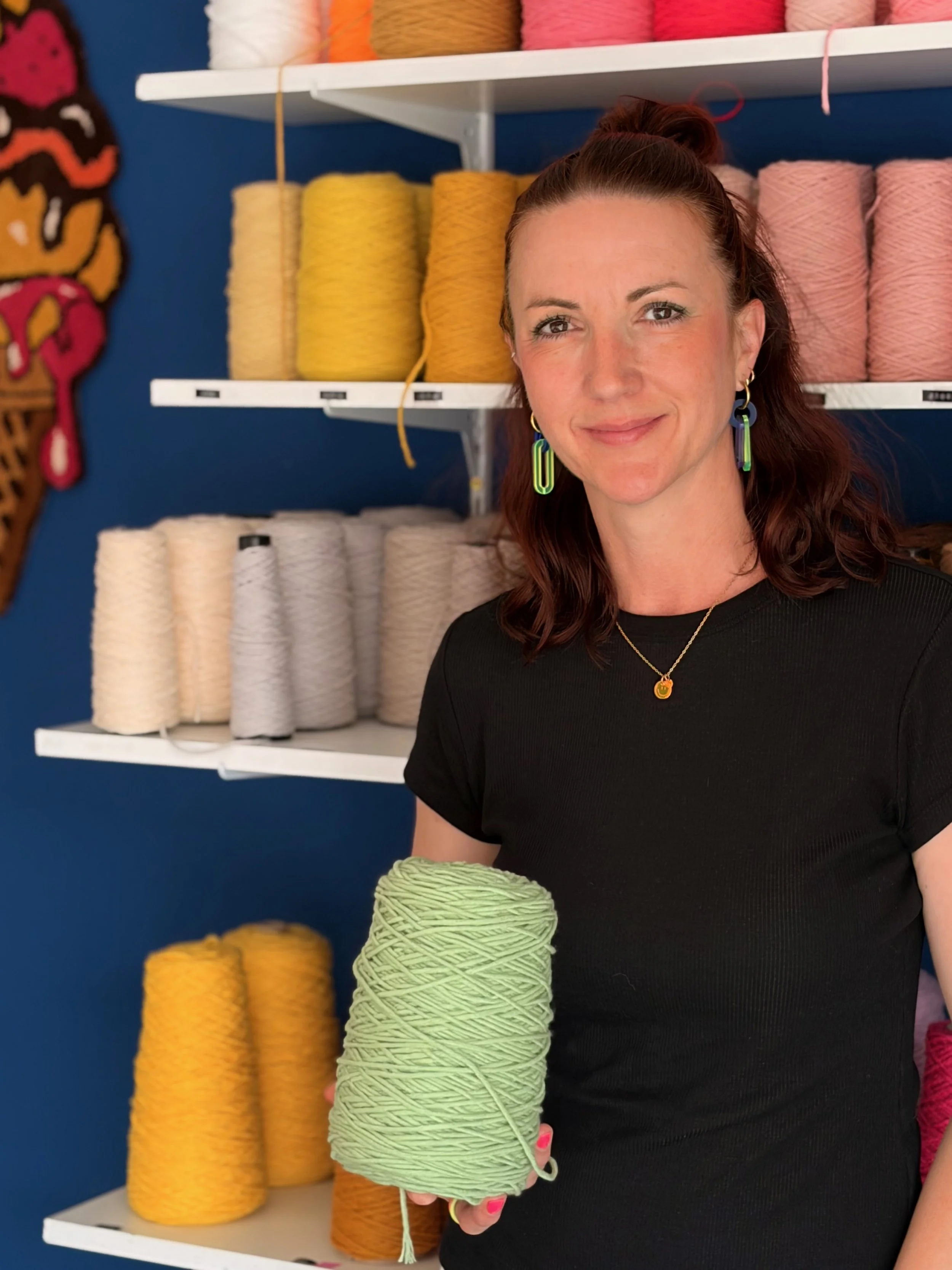 A woman holding a ball of light green yarn in a yarn store with shelves of colorful yarn in the background.
