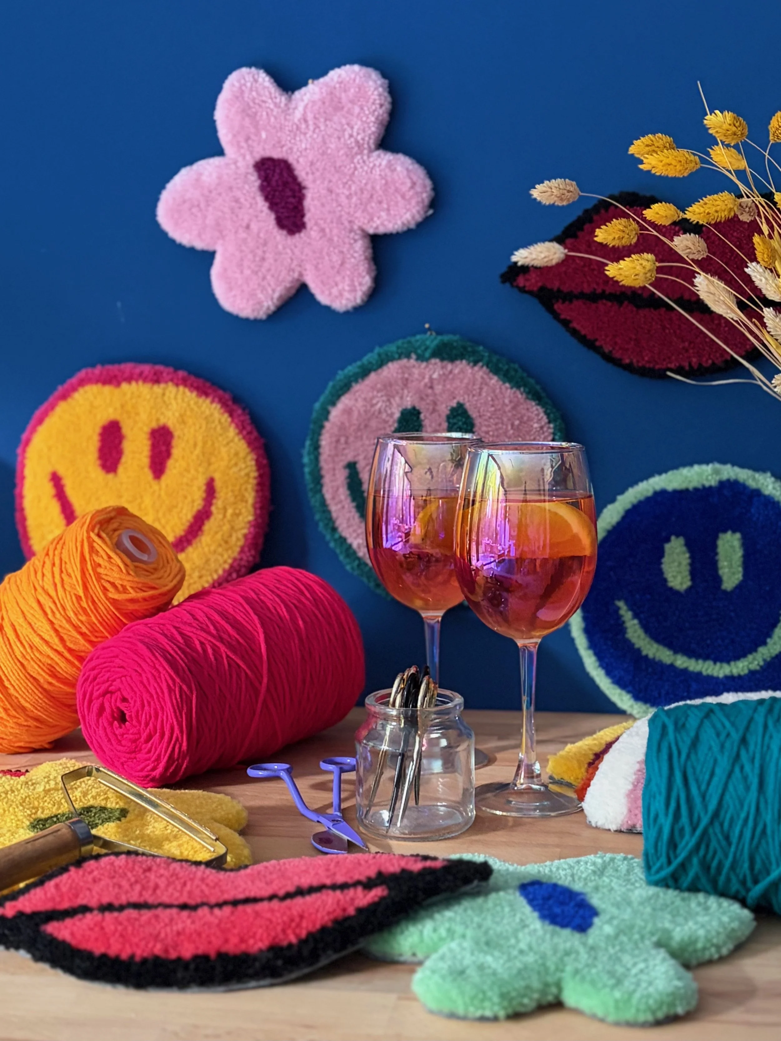 A colorful display of yarn in orange, pink, teal, and yellow, along with various felt and knitted items like smiley face patches, flowers, lips, and a star, arranged on a wooden surface with two glasses of rosé wine and a small jar of metal clips.