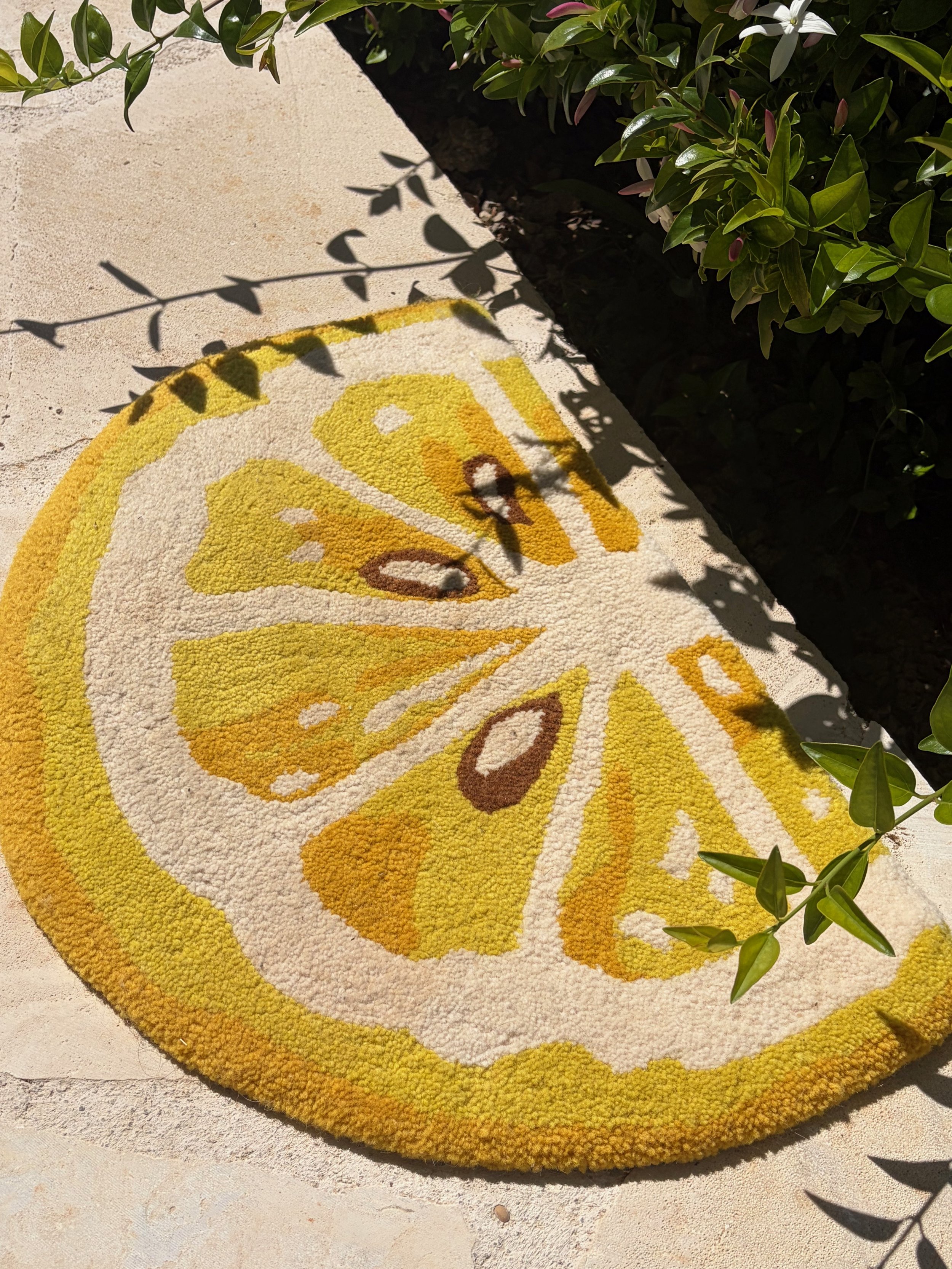 A yellow and white decorative outdoor rug with a butterfly design, placed on a light-colored concrete surface next to green bushes casting shadows.