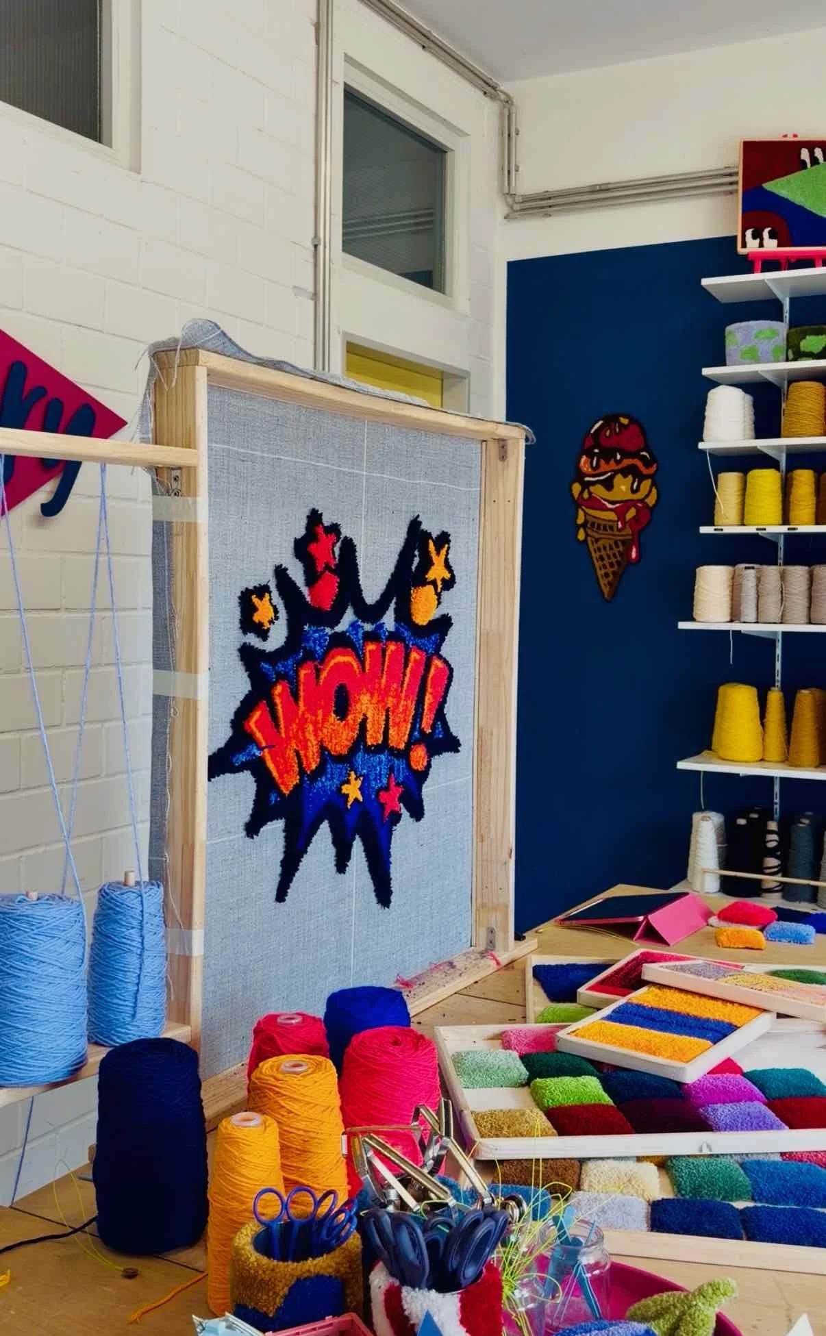 A crafting or art studio featuring yarn, colorful felt pieces, and a comic-style decoration on the wall that says "WOW!" The room has shelves with yarn in various colors, and a table with crafting supplies.
